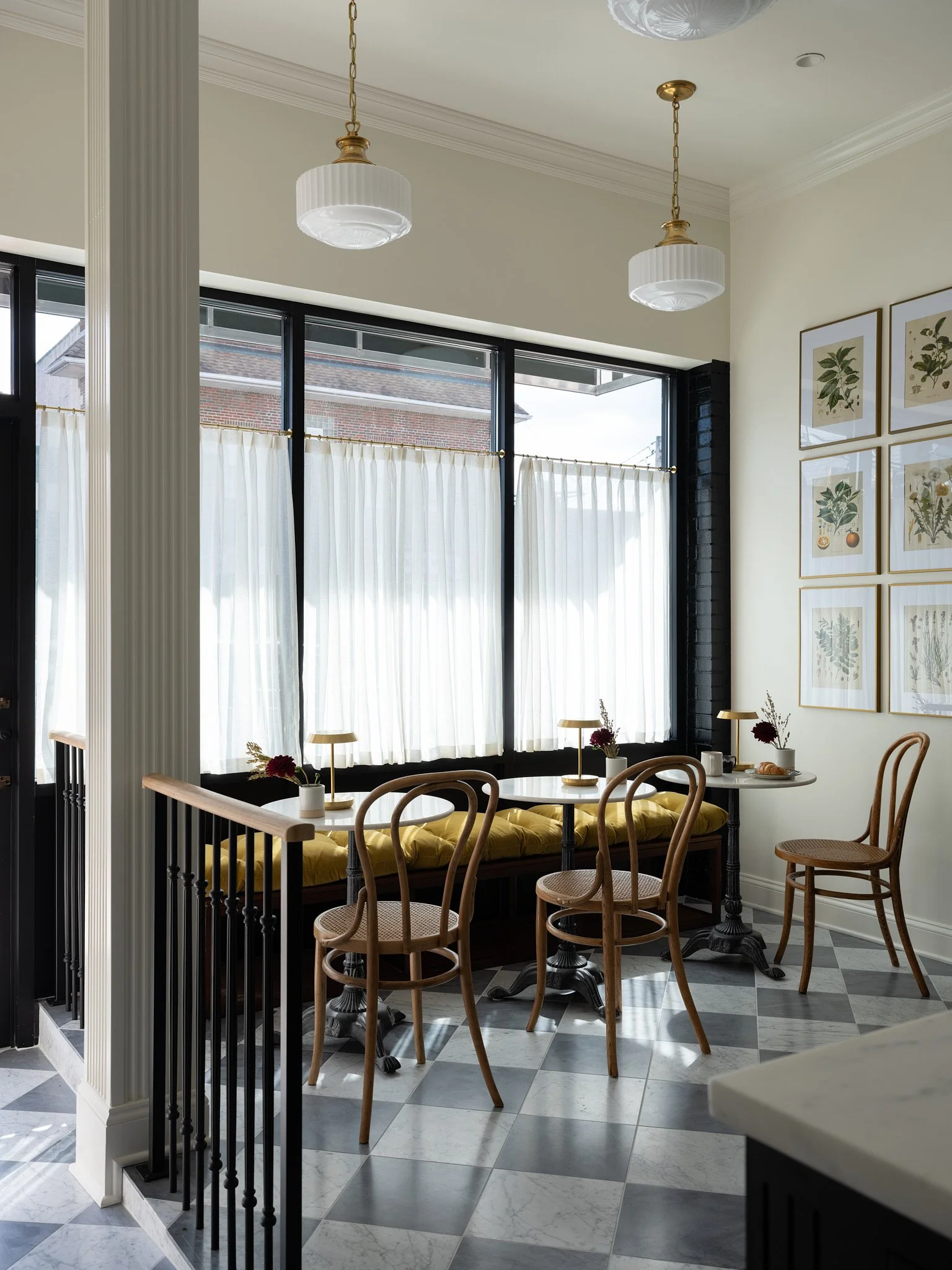 Interior of a cozy cafe with large windows, sheer white curtains, wooden chairs, small tables with lamps and vases, and framed botanical art on the wall.