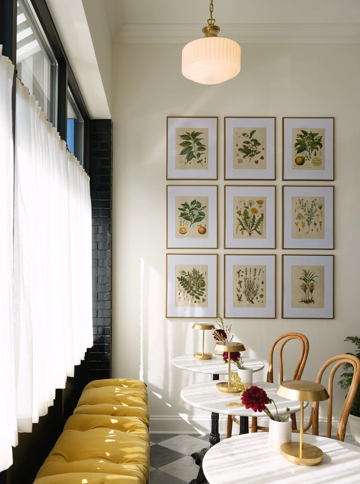 Interior of a bright room with a black brick wall, white curtains, a yellow tufted bench, round white marble tables with gold lamps and flower vases, wooden chairs, and framed botanical prints on the wall.