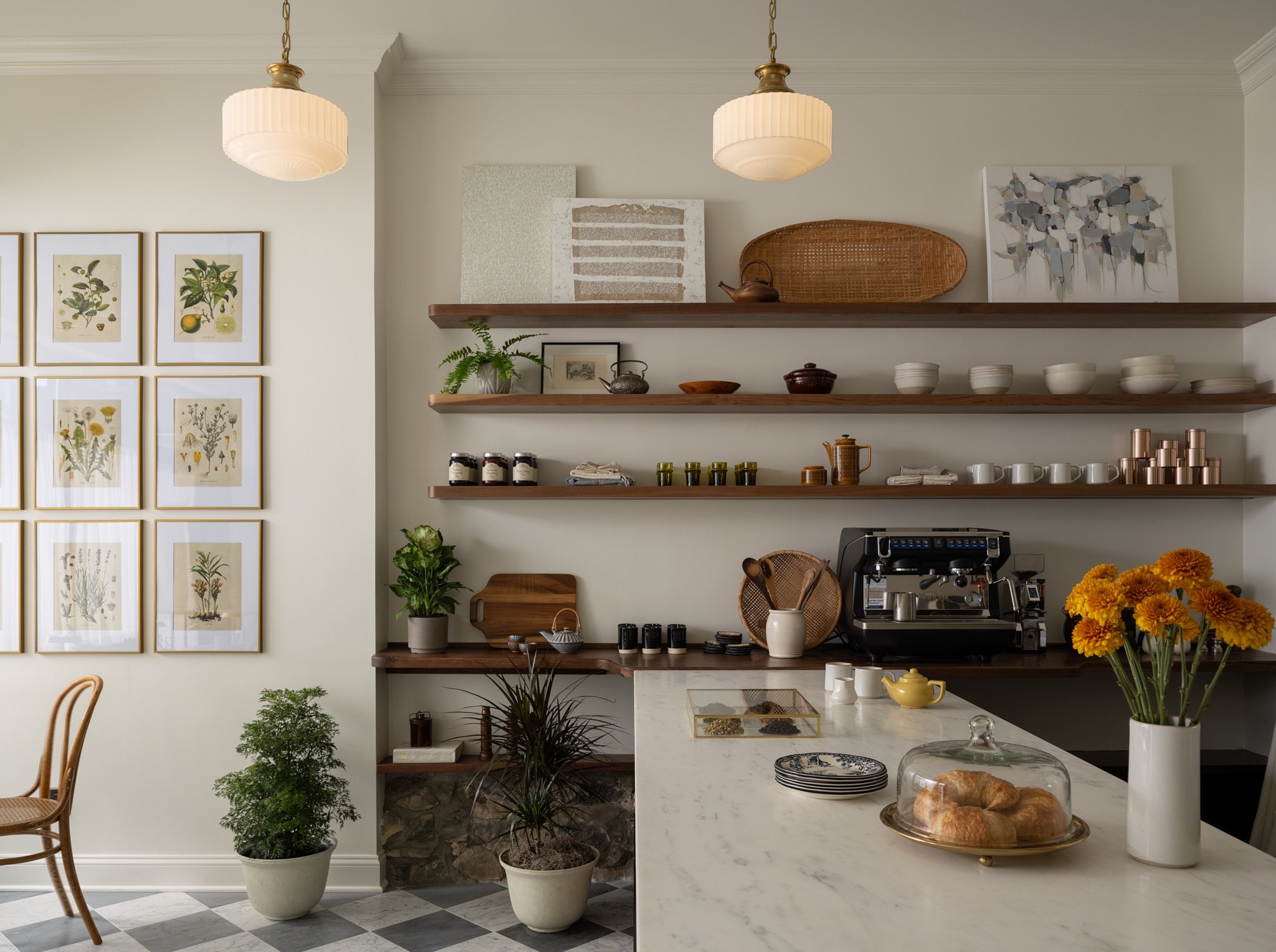 A cozy kitchen with open wooden shelves holding bowls, teapots, and cups. A marble counter with plates, a teapot, croissants under a glass dome, and a vase of orange flowers. Potted plants near a wall decorated with botanical prints. Two pendant ligh