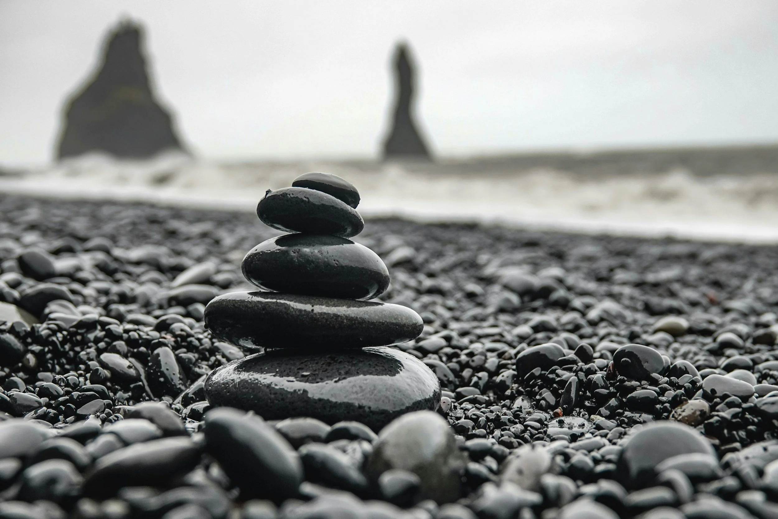 A stack of five smooth black stones on a pebble-covered beach with ocean waves and two large rock formations in the distance under an overcast sky.