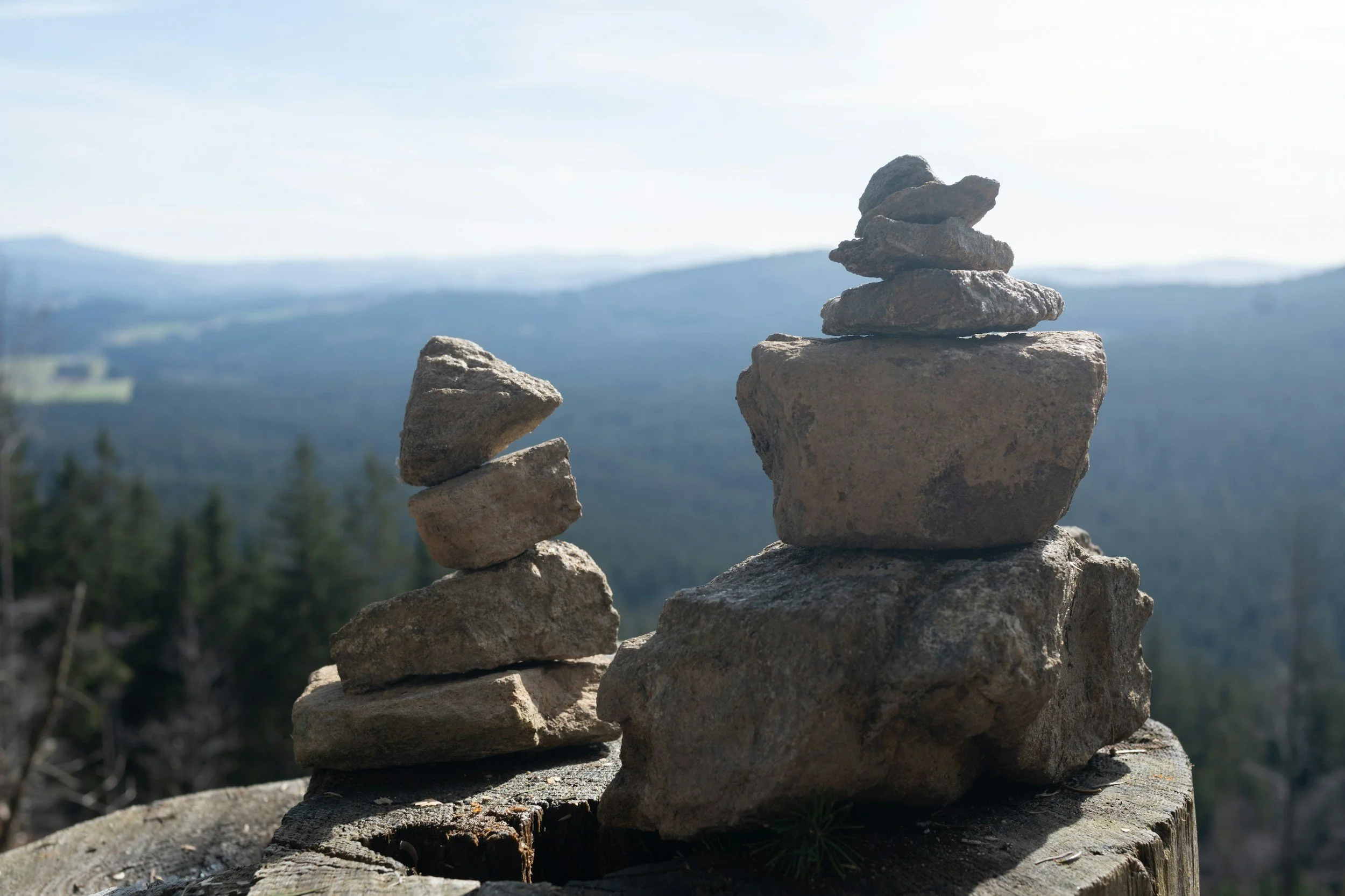 Two stacks of rocks, one larger and the other smaller, are placed on a wooden surface overlooking a forested valley with distant mountains and a partly cloudy sky in the background.