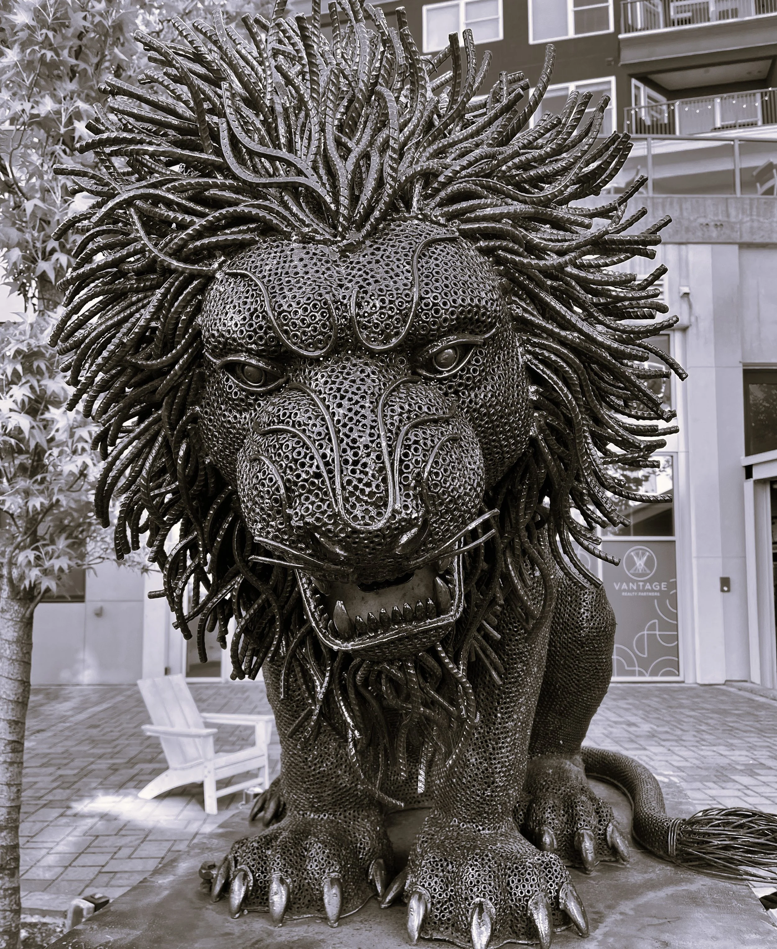 A large sculpture of a lion made from metal, with intricate details and a fierce expression, located outdoors in a city area.