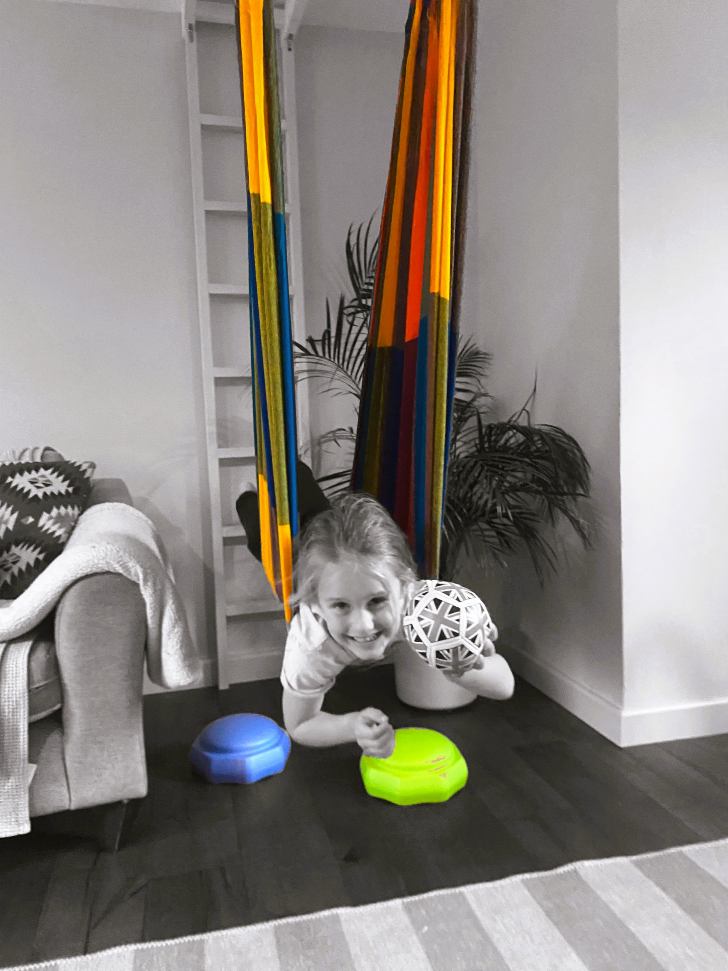 A young girl is lying on the floor, smiling at the camera, surrounded by colorful balance toys. The image is partially in black and white, with the toys in bright colors. There is a colorful hanging decorative piece and a potted plant in the background.