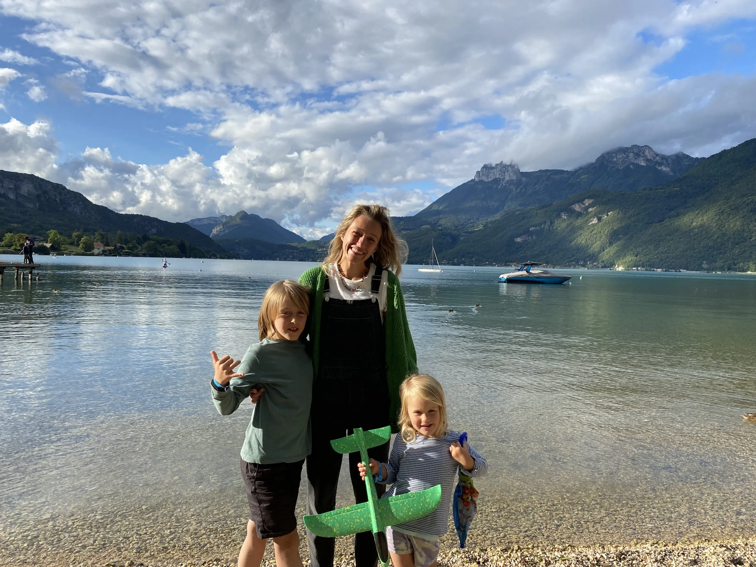 A woman with two children standing by a lake with mountains in the background. The woman has blonde hair and is wearing a green sweater and black overalls. One child has blonde hair and is making a hand gesture, the other child has light brown hair and is holding a toy-shaped object. There are boats on the water, and the sky is partly cloudy.