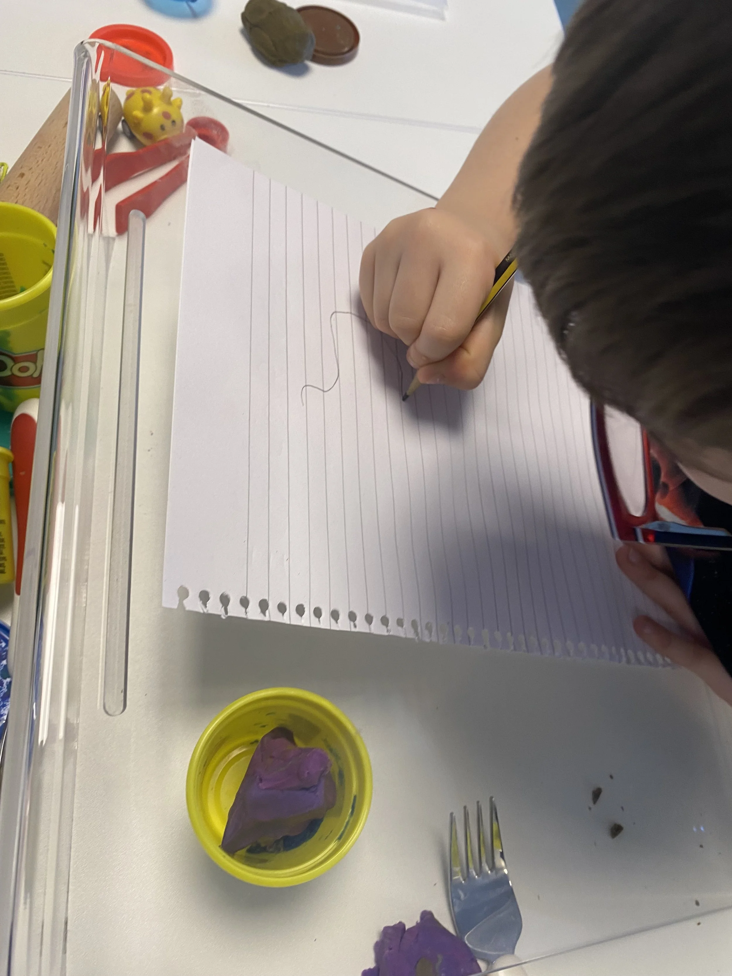 A child with brown hair is drawing on a lined sheet of paper with a pencil on a white table. There are small containers of playdough, including purple and yellow, and some chopped pieces of playdough on the table. There is also a fork nearby and some scattered small bits of what appears to be dirt or crumbs.