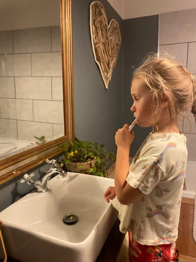 A young girl with light brown hair tied back, brushing her teeth in a bathroom. She wears a white T-shirt with rainbow patterns and red pajama pants. The bathroom has a white sink with silver faucets, a large mirror with a wooden frame, and some green plants. There is a heart-shaped wall hanging above the sink.