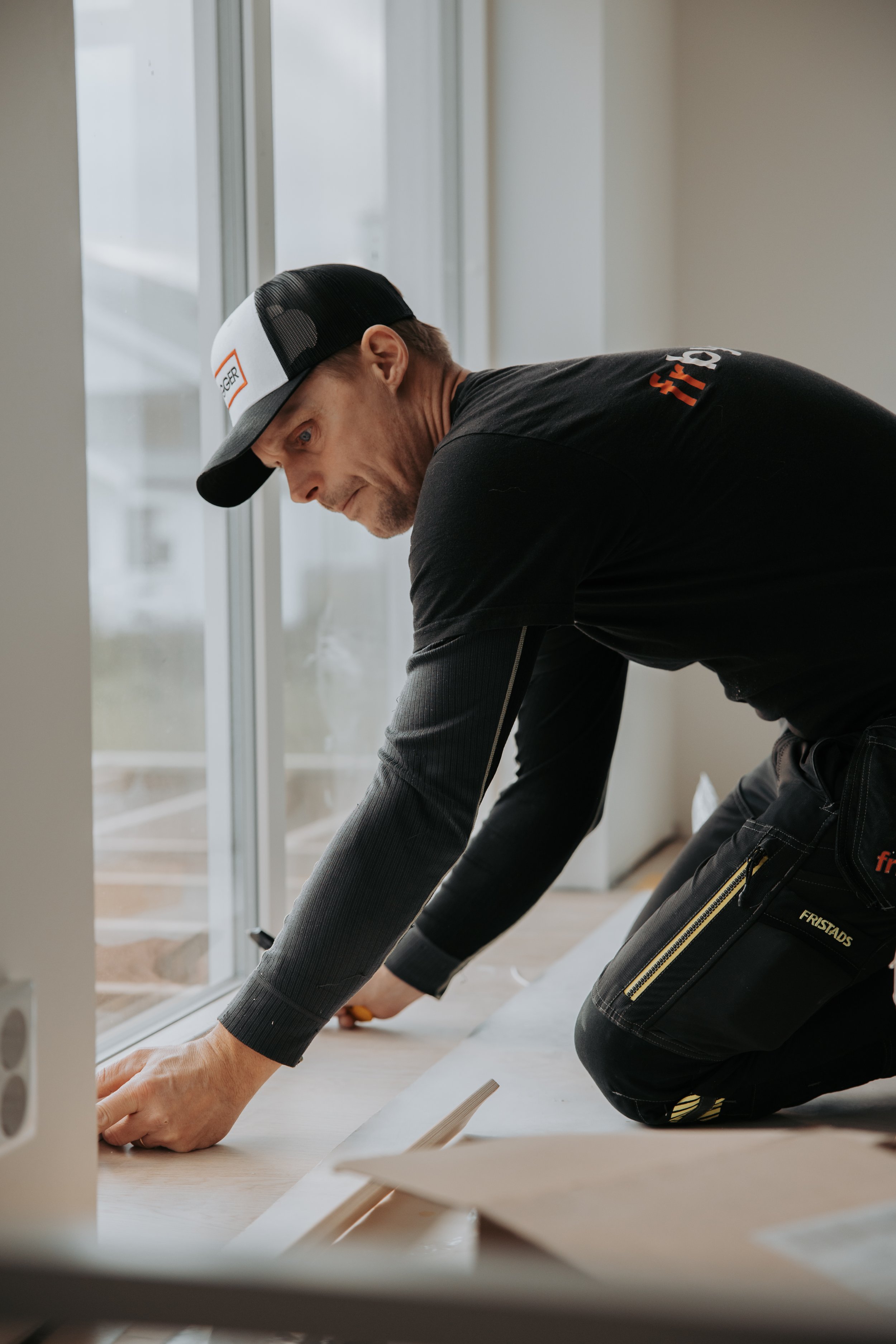 En man som arbetar med att installera golv, kröker sig på knä på ett rum med stora fönster.