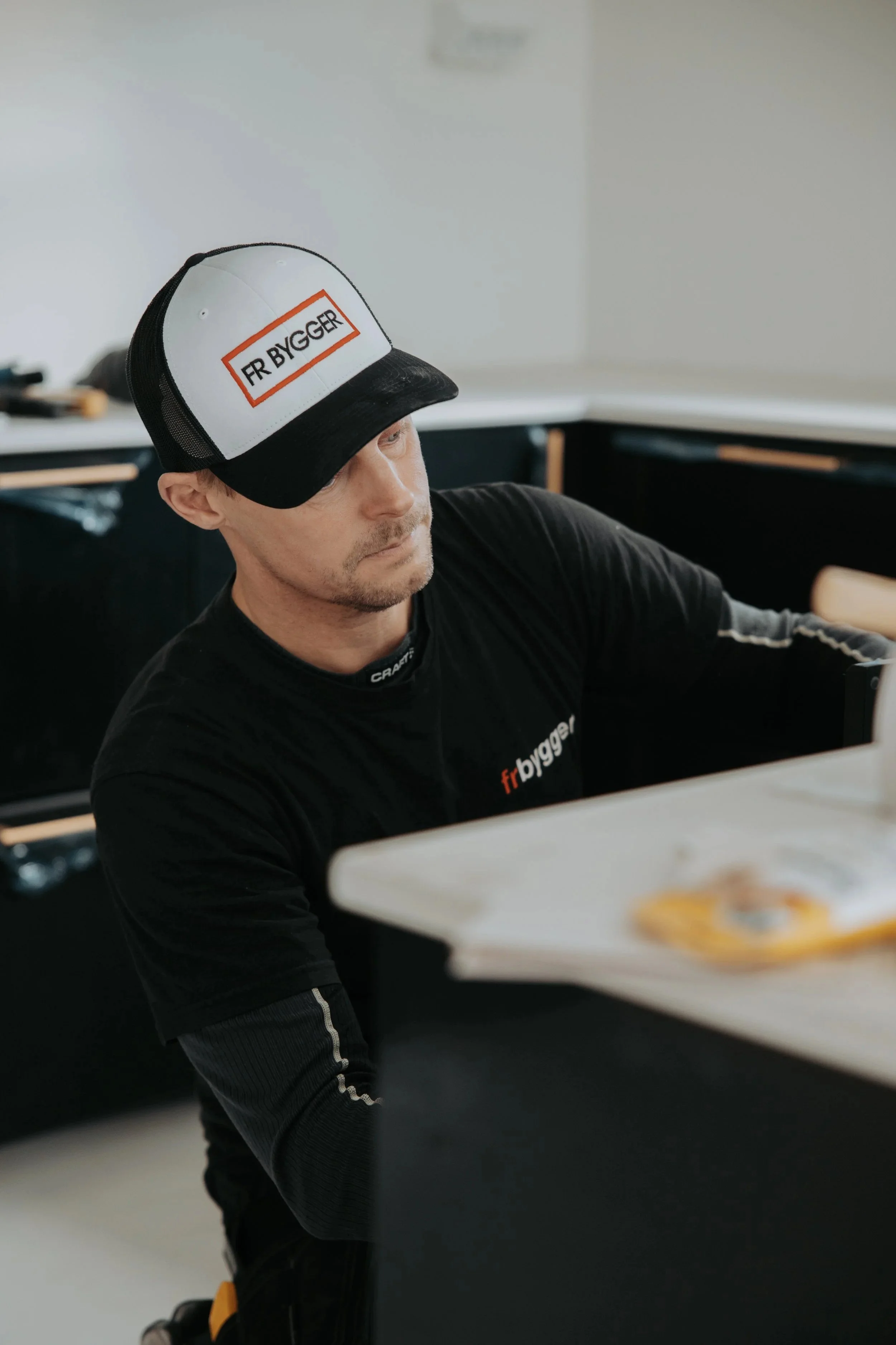 En man med svart tröja och en svart och vit trucker keps med texten 'FR BYGGER' under ett arbetsbord