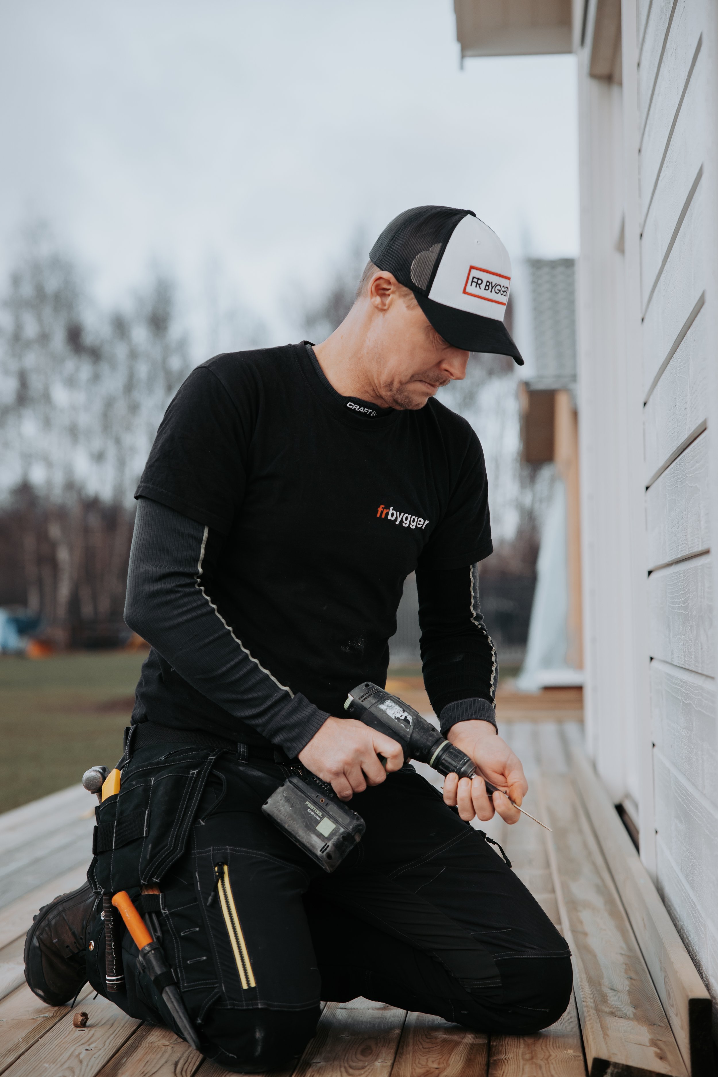 En man i svart tröja och keps arbetar med en skruvmejsel vid sidan av ett hus.