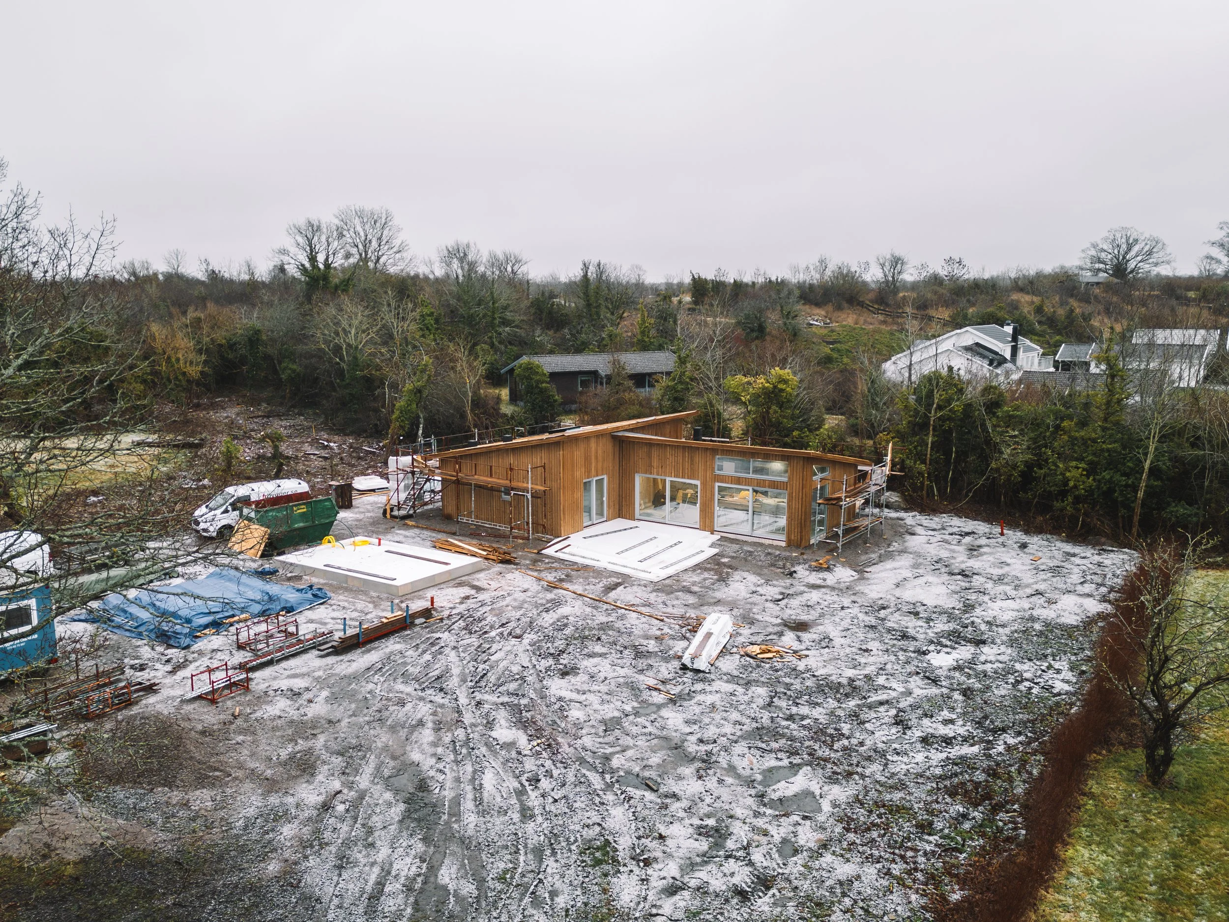 Byggarbetsplats med ett nybyggt hus av trä, omgiven av förberedelser för ytterligare byggnation, med några byggmaterial, fordon och utrustning på plats.