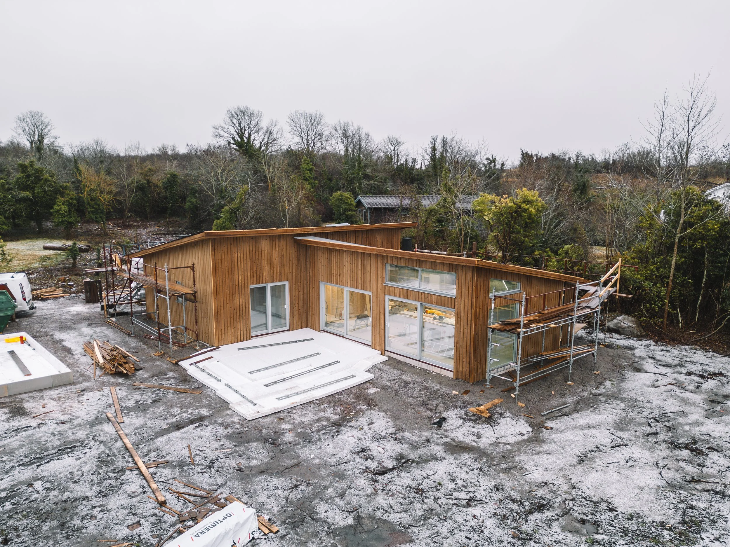 En pågående byggarbetsplats med ett hus under konstruktion. Huset har träväggar och stora fönster. Runt byggnaden finns byggnadsställningar och byggmaterial. Marken är ojämn och delvis snöbelagd, och det finns träd i bakgrunden.