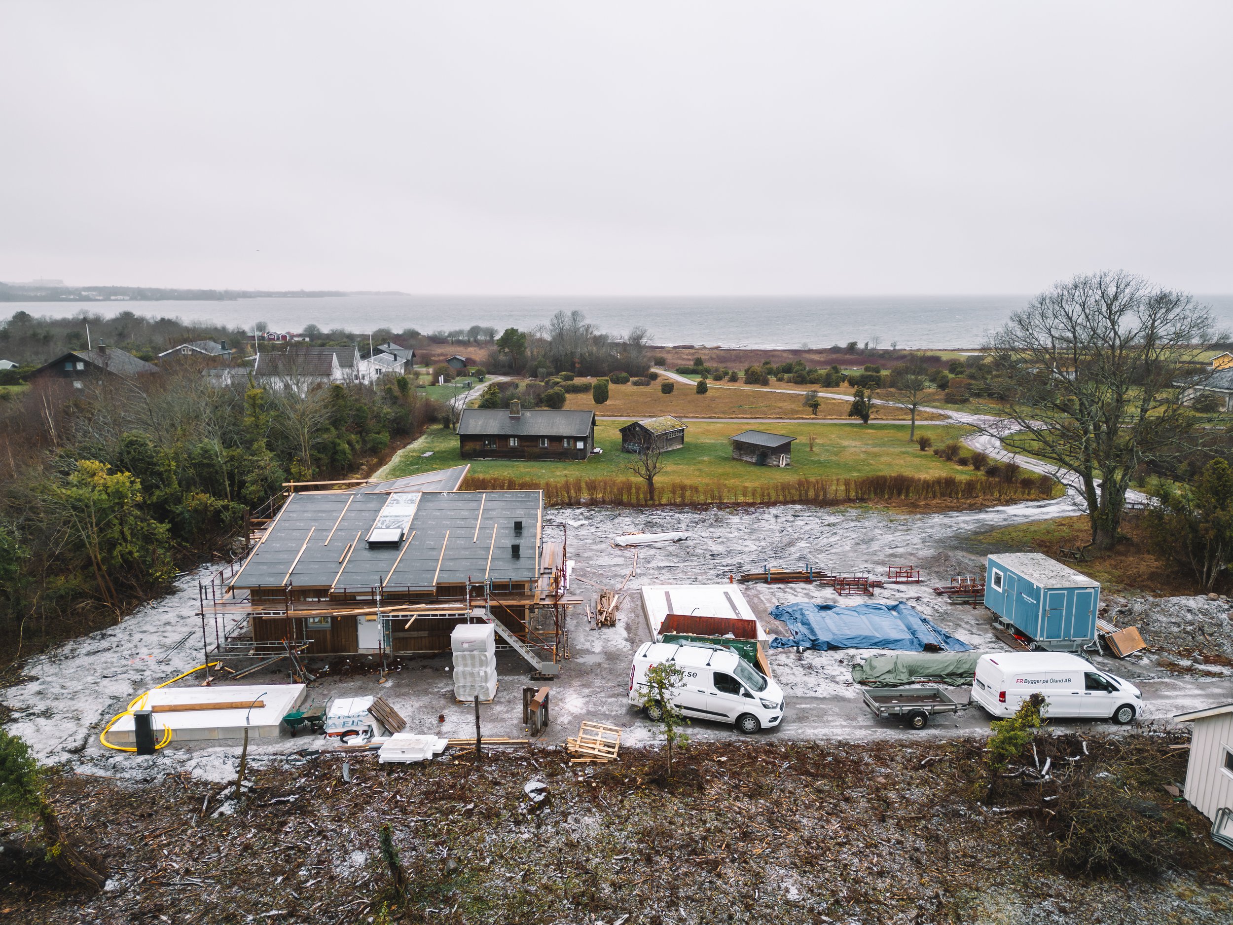 Byggnation av ett hus vid kusten med tillfälliga byggmaterial, bilar och utomhusutrustning i förgrunden.
