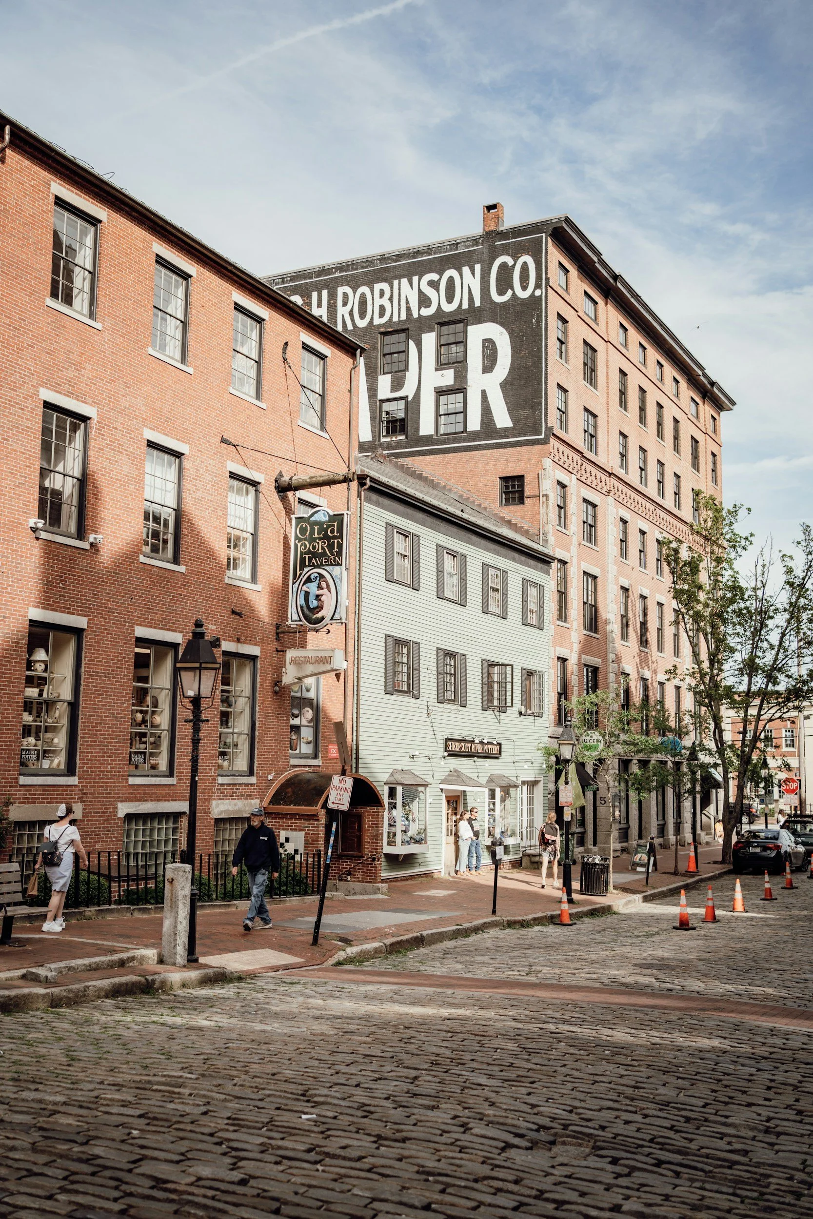 Street scene with red brick buildings and a large painted building in the background advertising 'H. Robinson Co. Pier'. There are pedestrians walking, street lamps, parking cones, and parked cars along the street.