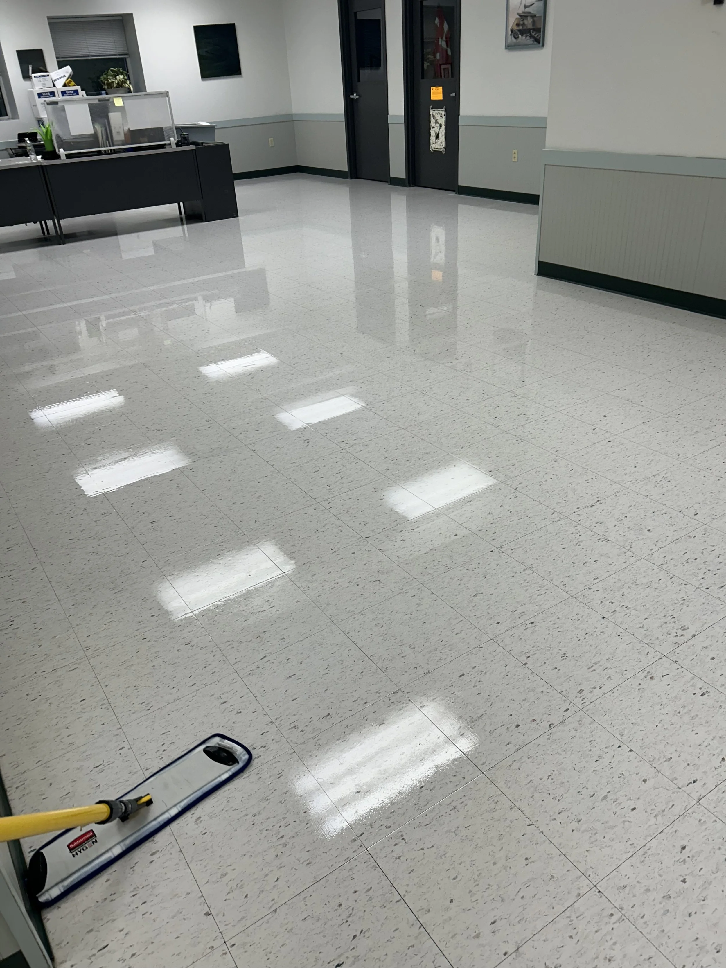 Empty office or lobby with shiny tiled floor, a mop and bucket in the foreground, a counter with office supplies, and closed door in the background.