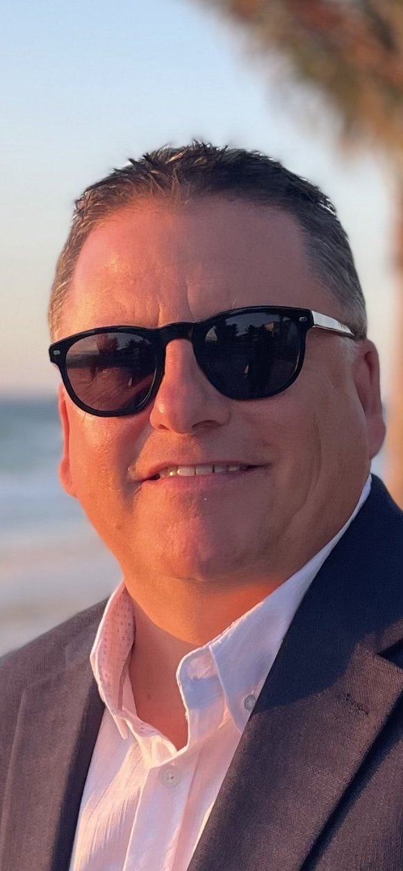 A man wearing sunglasses and a white shirt with a dark jacket, standing outdoors near the beach during sunset.