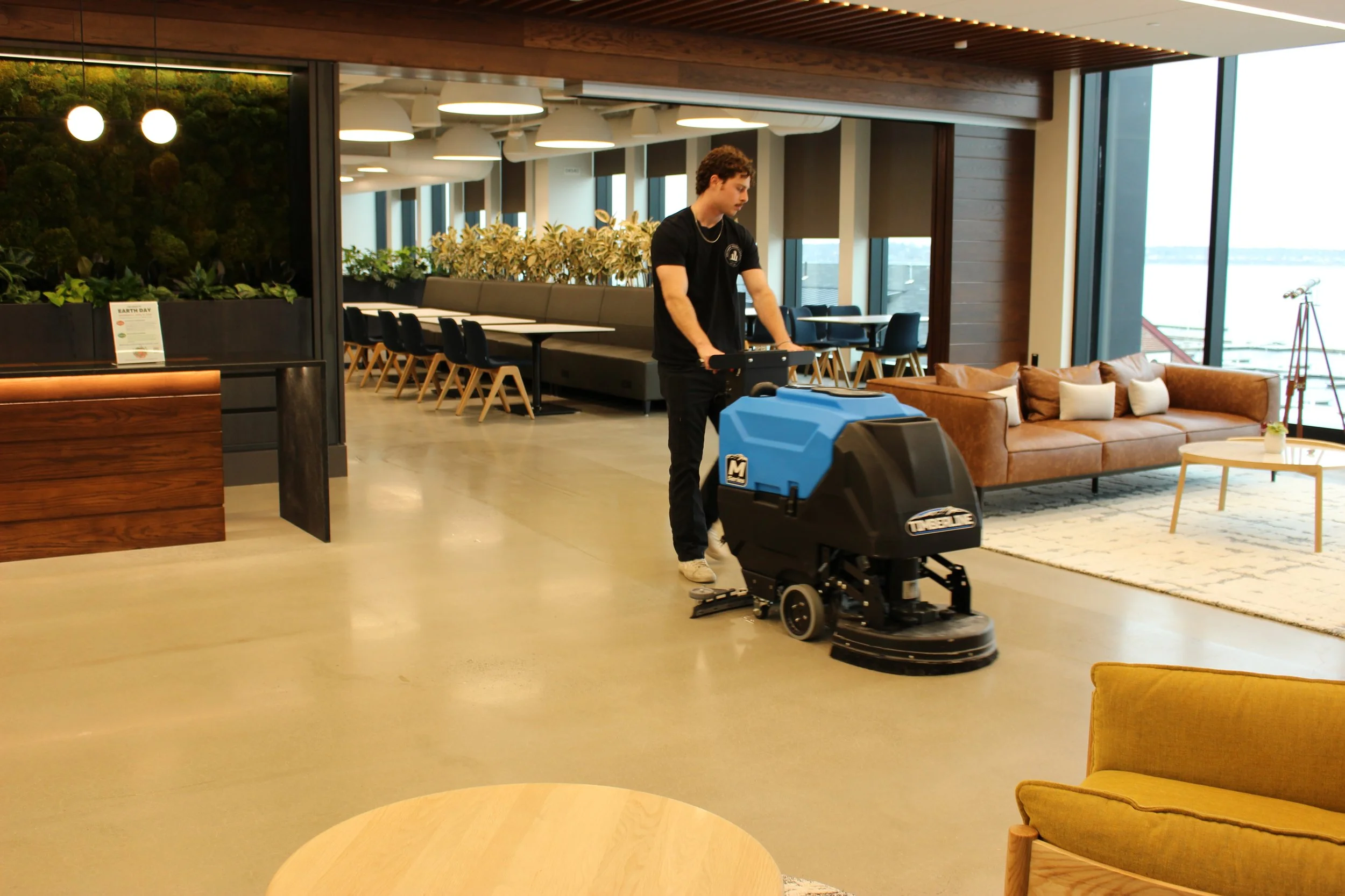 A person is cleaning a large open indoor space with a floor cleaning machine. The space appears to be a modern lounge or waiting area with tables, chairs, couches, and large windows showing an outside view.