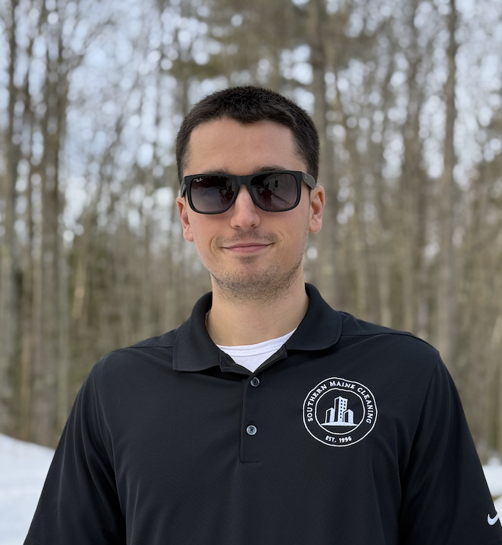 A young man with short dark hair wearing sunglasses, a black collared shirt with a logo, standing outdoors in a wooded area with snow on the ground.