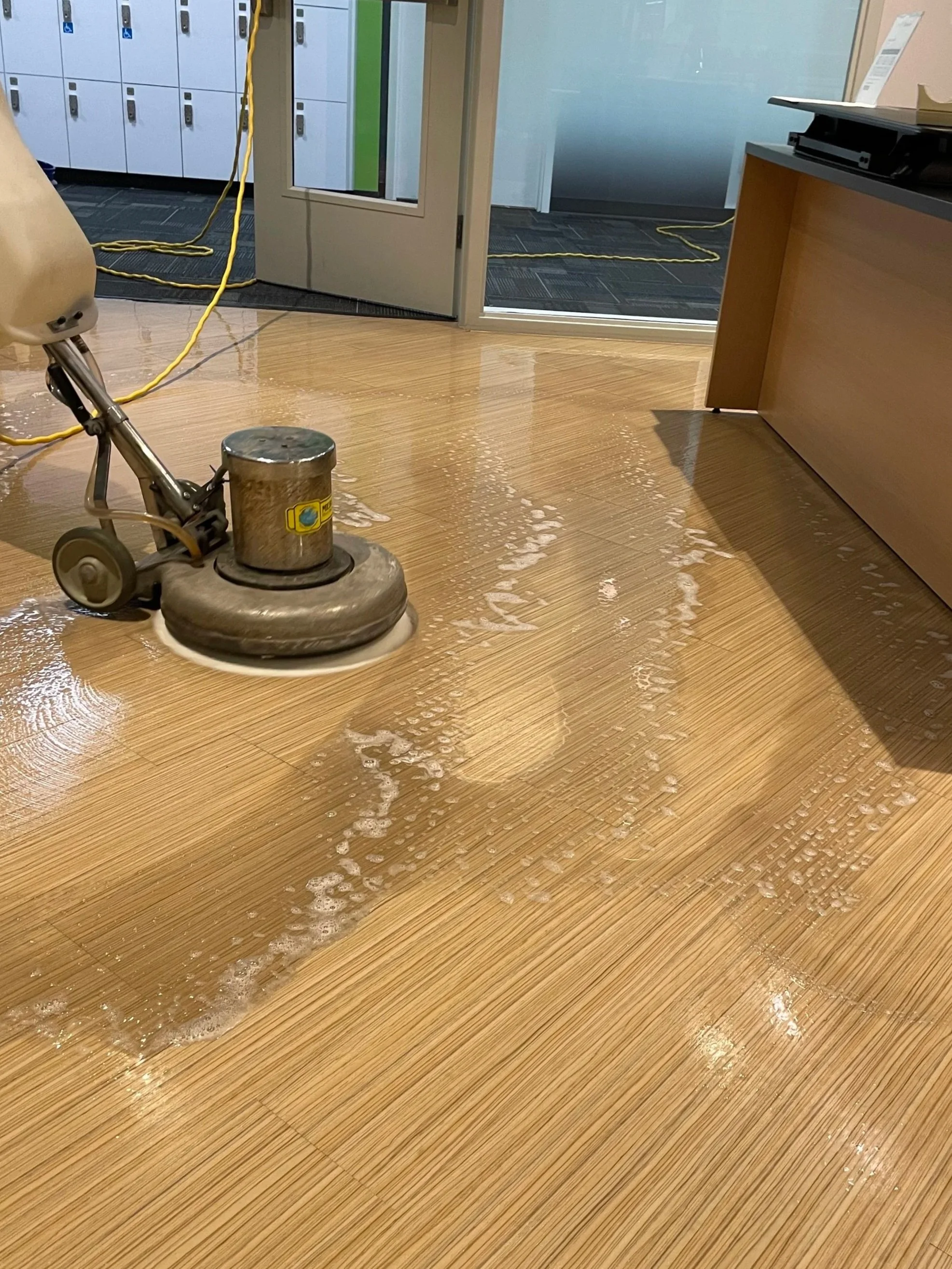 A floor polishing machine is in use, cleaning and shining a wood floor inside a building. There are lockers in the background, and some furniture and office supplies are visible.