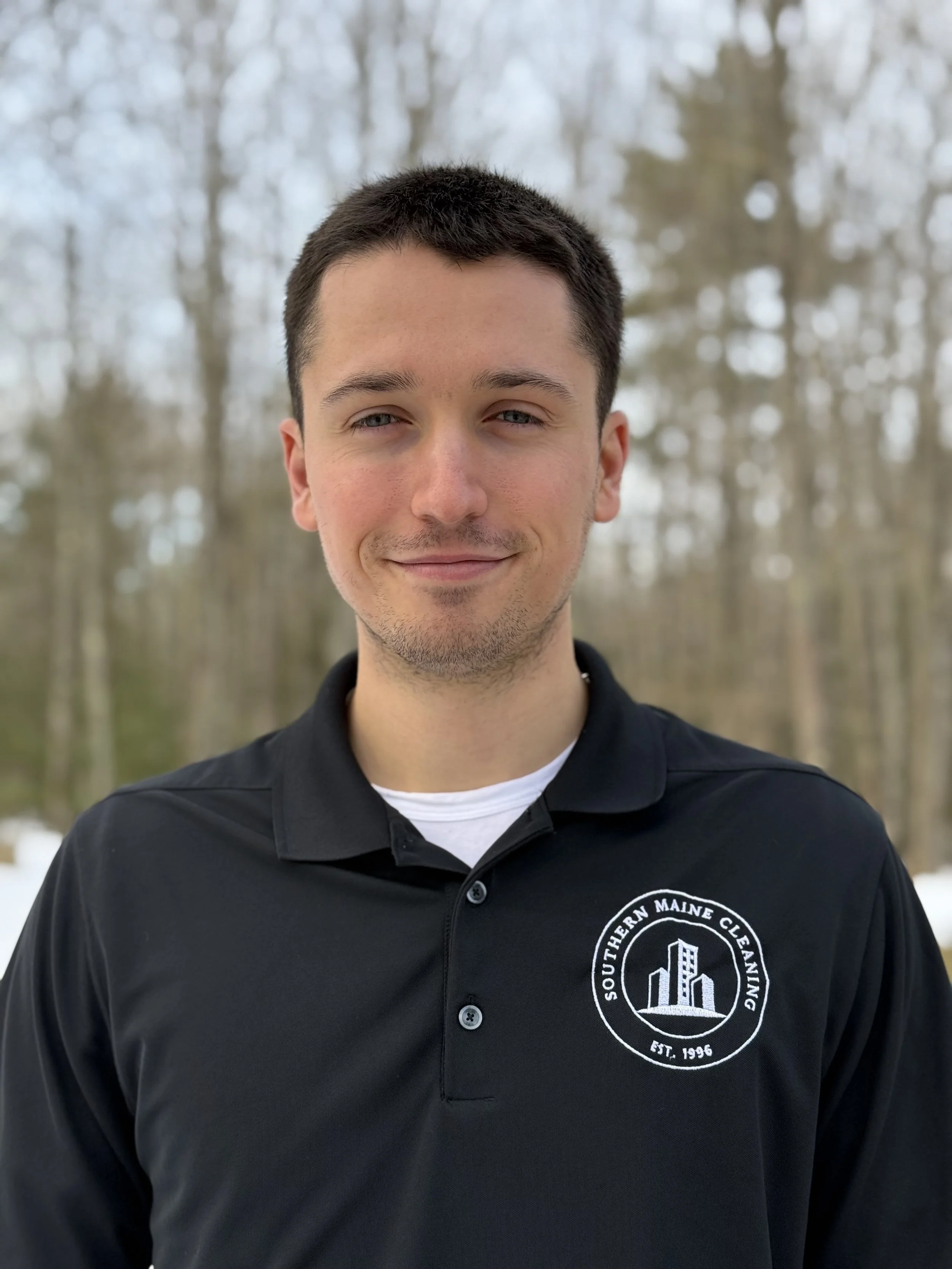 A young man with short dark hair, wearing a black polo shirt with a logo on the chest that reads 'Southern Maine Cleaning' and features an image of buildings, standing outdoors in front of a blurred background of trees with no leaves.