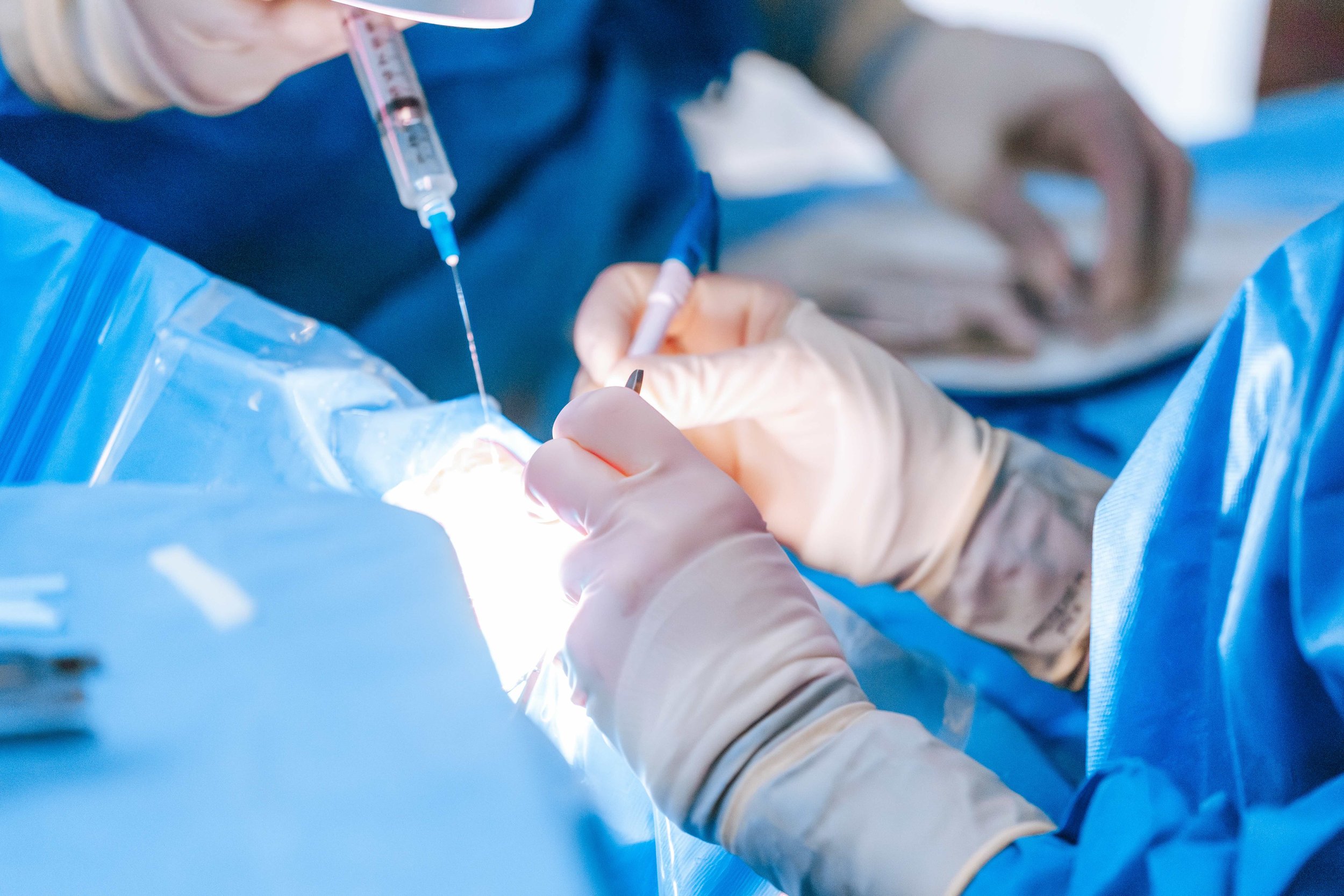 Surgeons in blue gowns and gloves performing a glaucoma operation with surgical tools and a syringe under bright lighting.