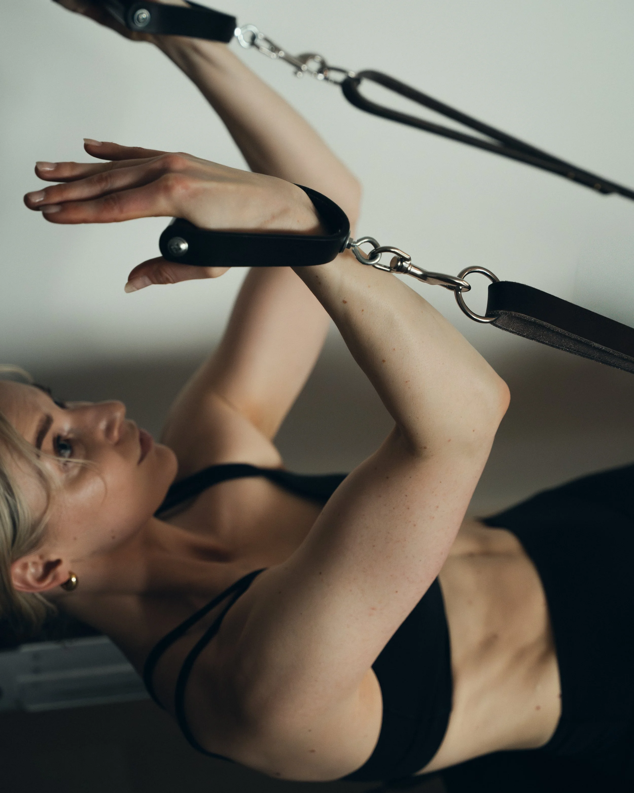 A woman with blonde hair and fair skin lying on her back, looking at her hands, with her arms raised. She is wearing a black lingerie top with thin straps and a black bracelet on her wrist. The photo captures her from a side angle, with a focus on he
