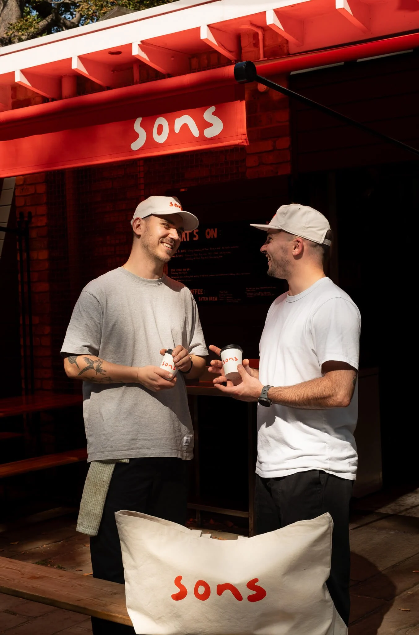 Two men smiling and talking to each other outside a restaurant, holding cups of coffee, with a red awning and a brick wall behind them. The awning and cups have the word "sons" written on them.