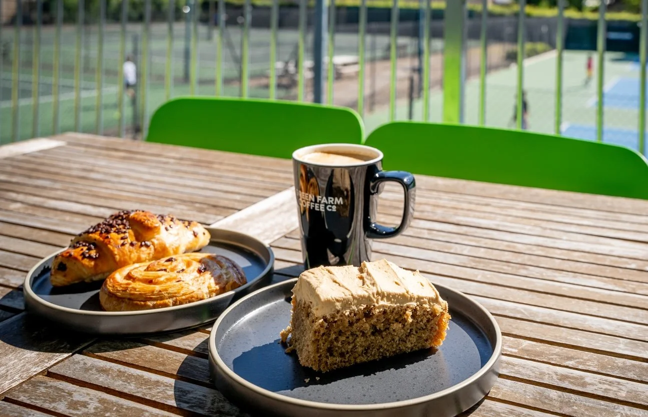 Coffee, cake and pastries on a table in the sunshine with the tennis courts behind them.