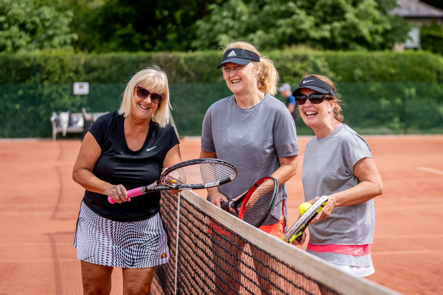 Three women on a tennis court enjoying a game, holding tennis rackets, smiling and laughing, dressed in athletic clothing and sunglasses, with a green fence and trees in the background.