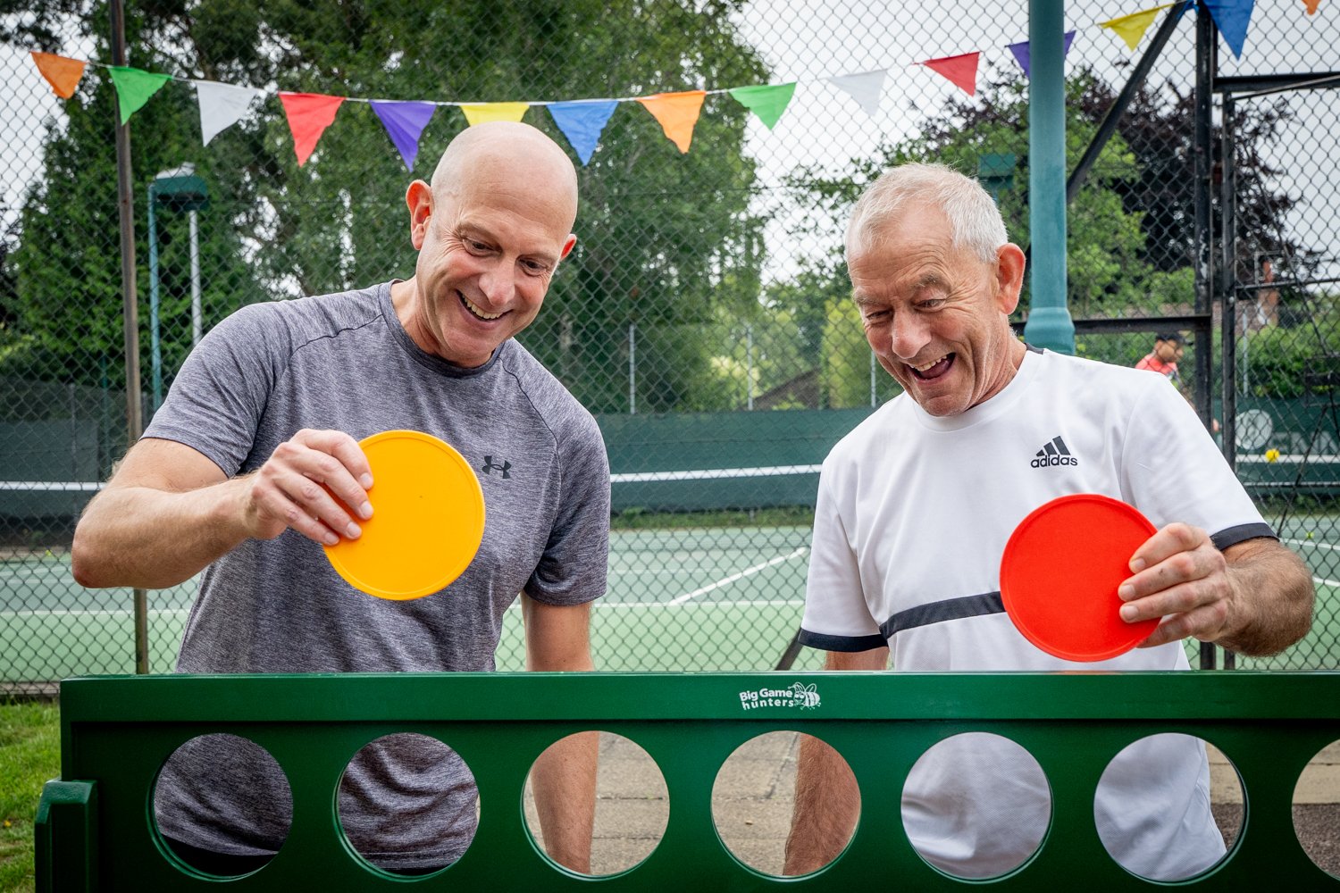 Two male members playing giant connect 4.  They are both smiling and laughing.  Their is a tennis court in the background with bunting on the fence.