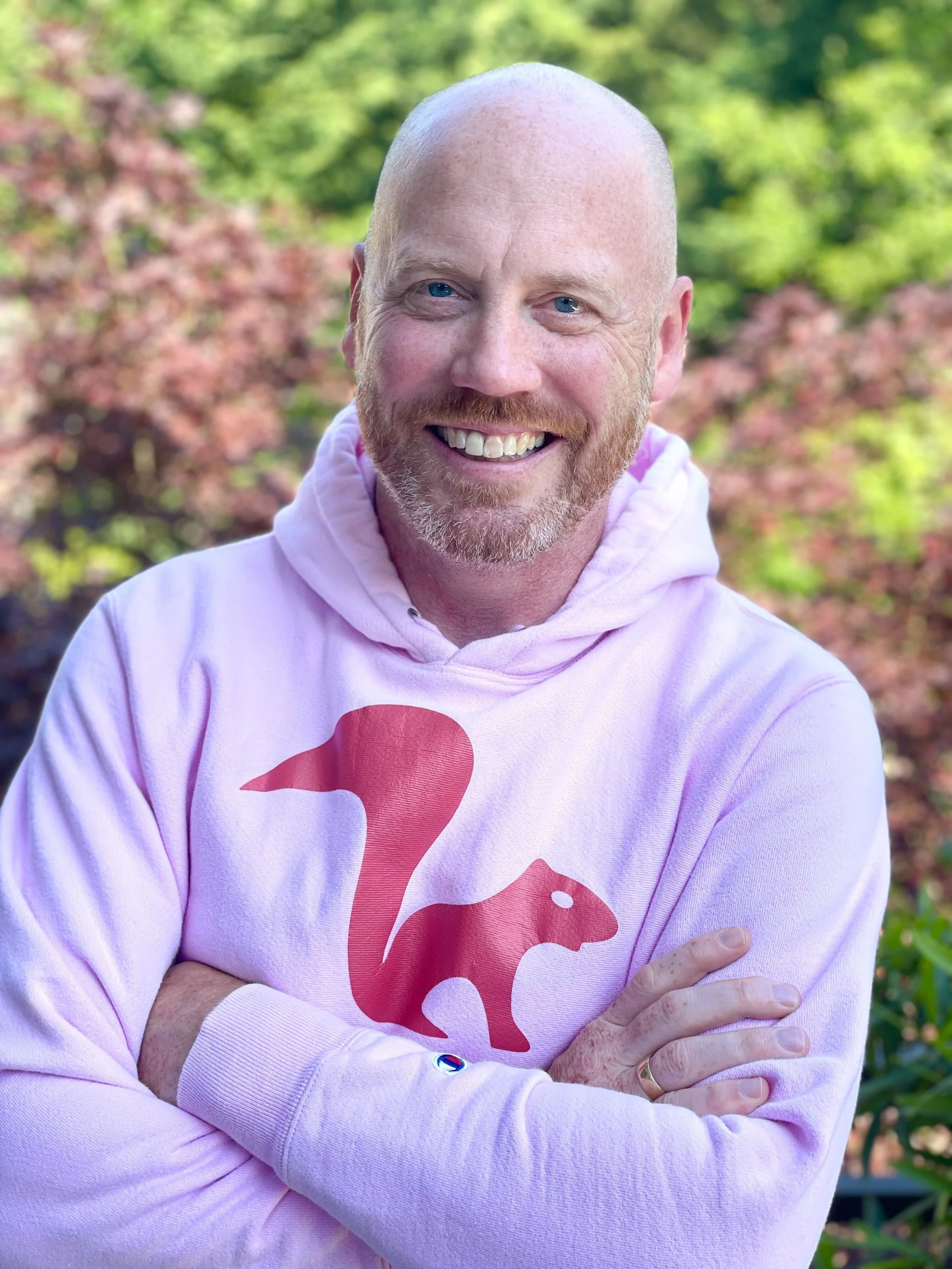 A smiling man with a bald head and blue eyes, wearing a light pink hoodie with a red squirrel logo on the front, standing outdoors with green and pink trees in the background.