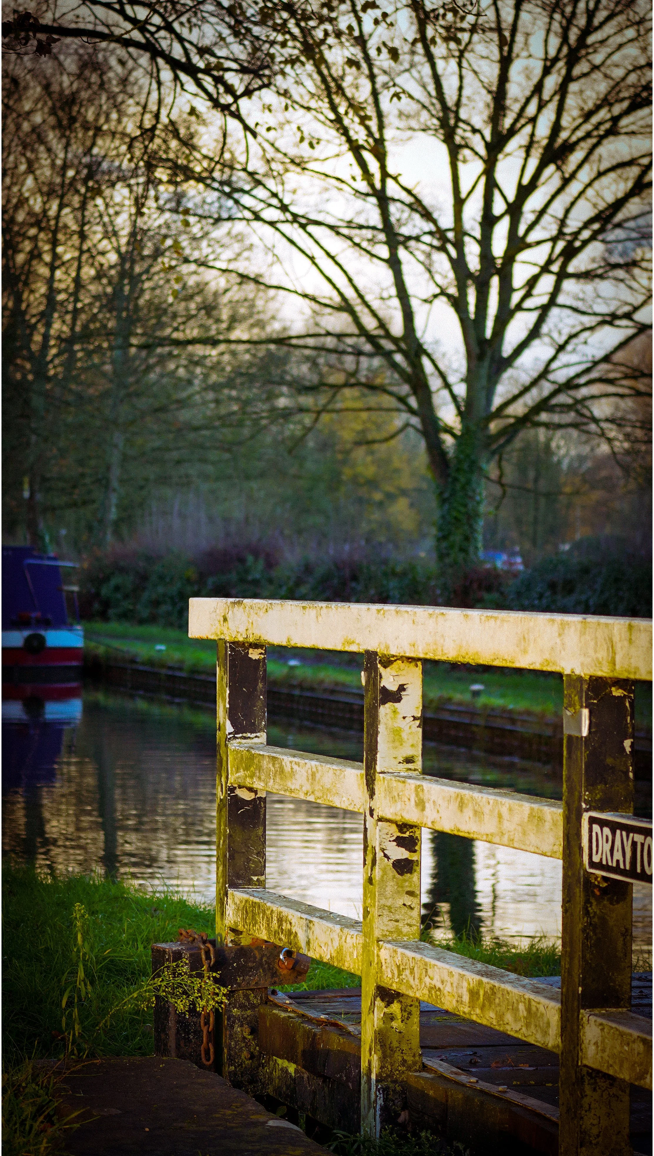 A weathered yellow and black metal railing along a canal with ships, trees, and a cloudy sky in the background.