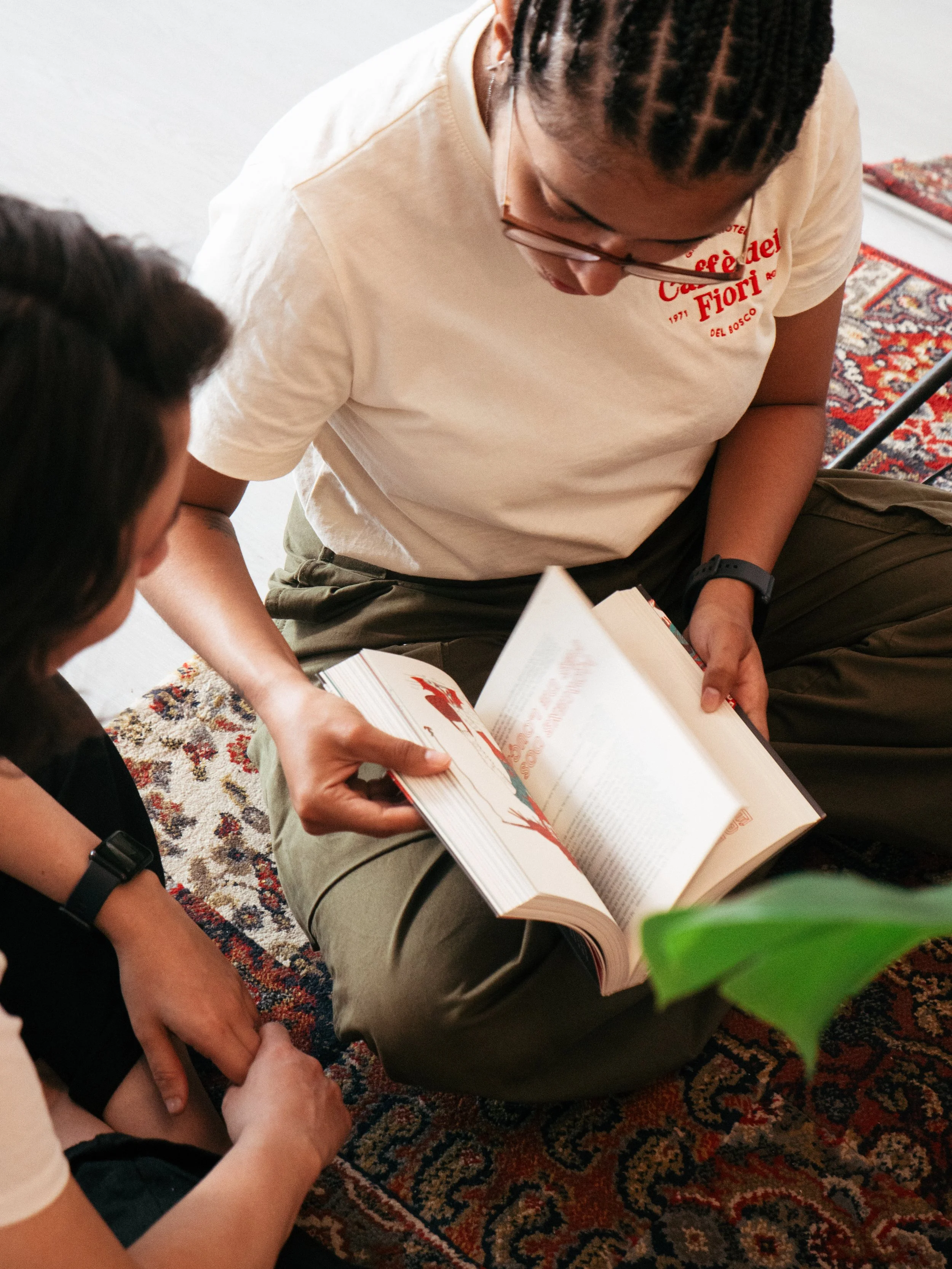 Pessoa com cabelo trançado, usando óculos e camiseta clara, sentada no chão, lendo um livro aberto para outra pessoa que também está sentada no chão ao lado, em um ambiente com tapete de padrão vibrante.