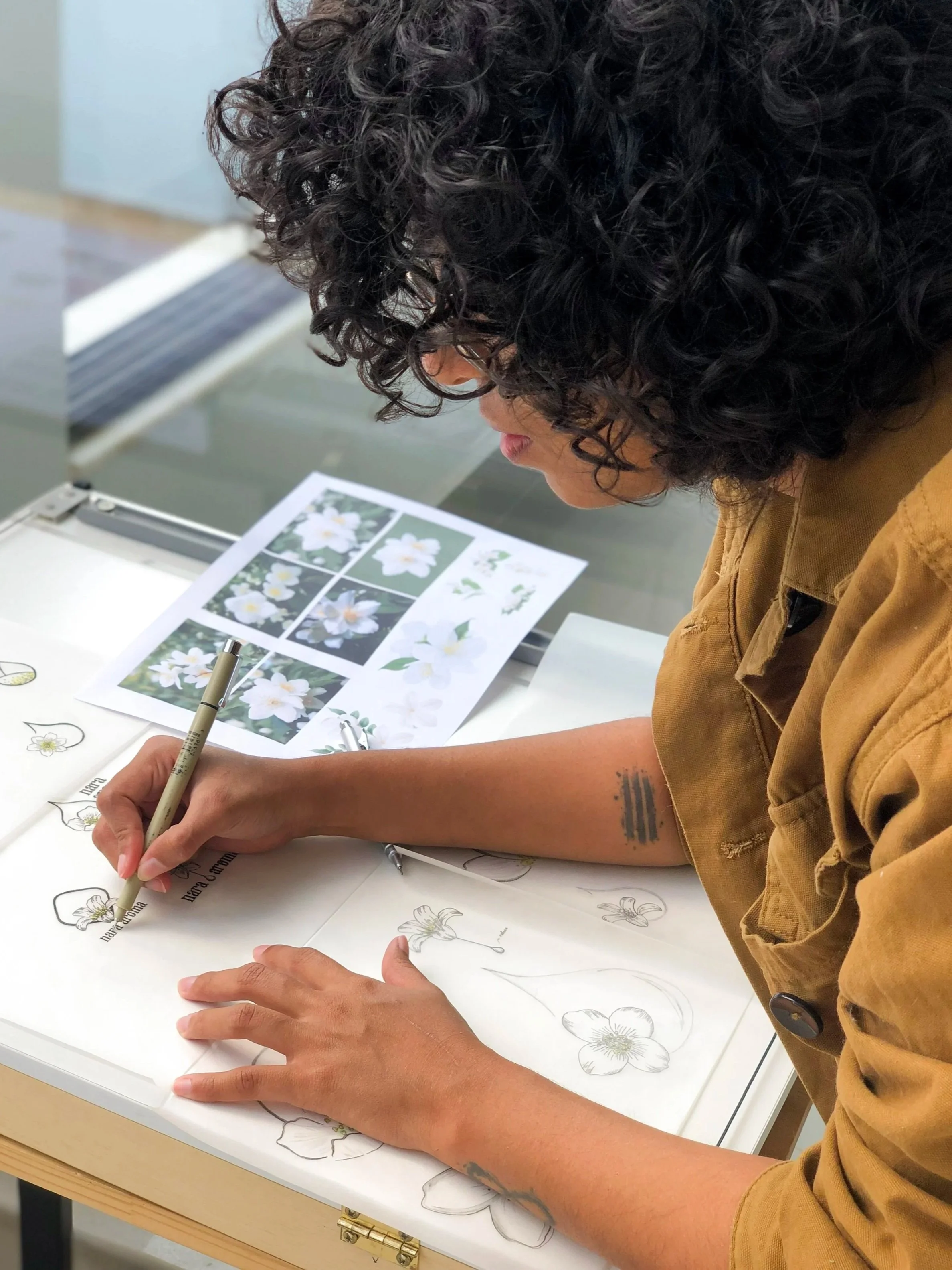 Pessoa com cabelo cacheado escuro desenhando flores em um papel, com referência às fotos de flores brancas na mesa.