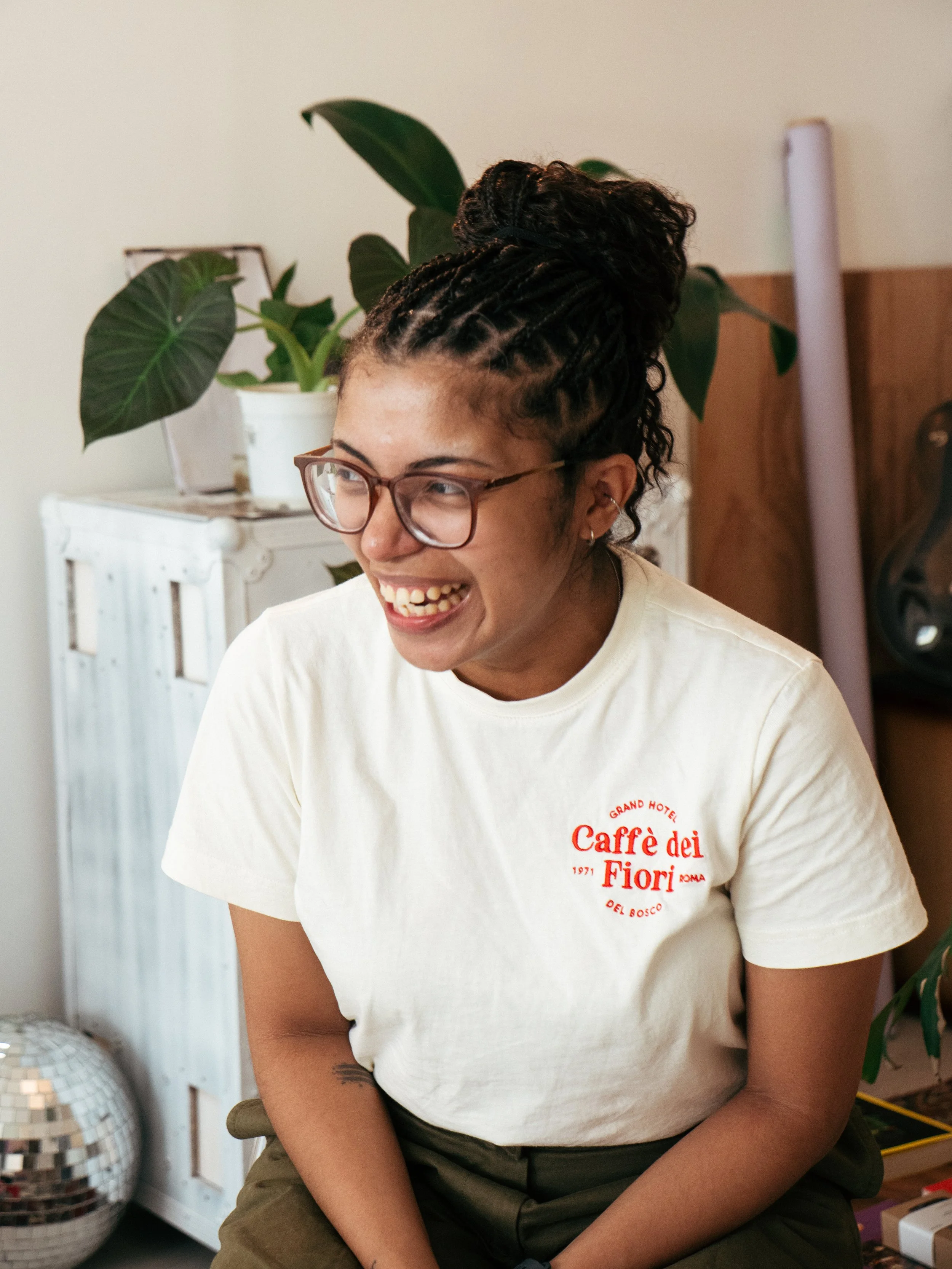 Mulher sorrindo, usando óculos, com cabelo em box braids e camiseta branca com logotipo vermelho, sentada em ambiente interior com plantas e objetos decorativos.