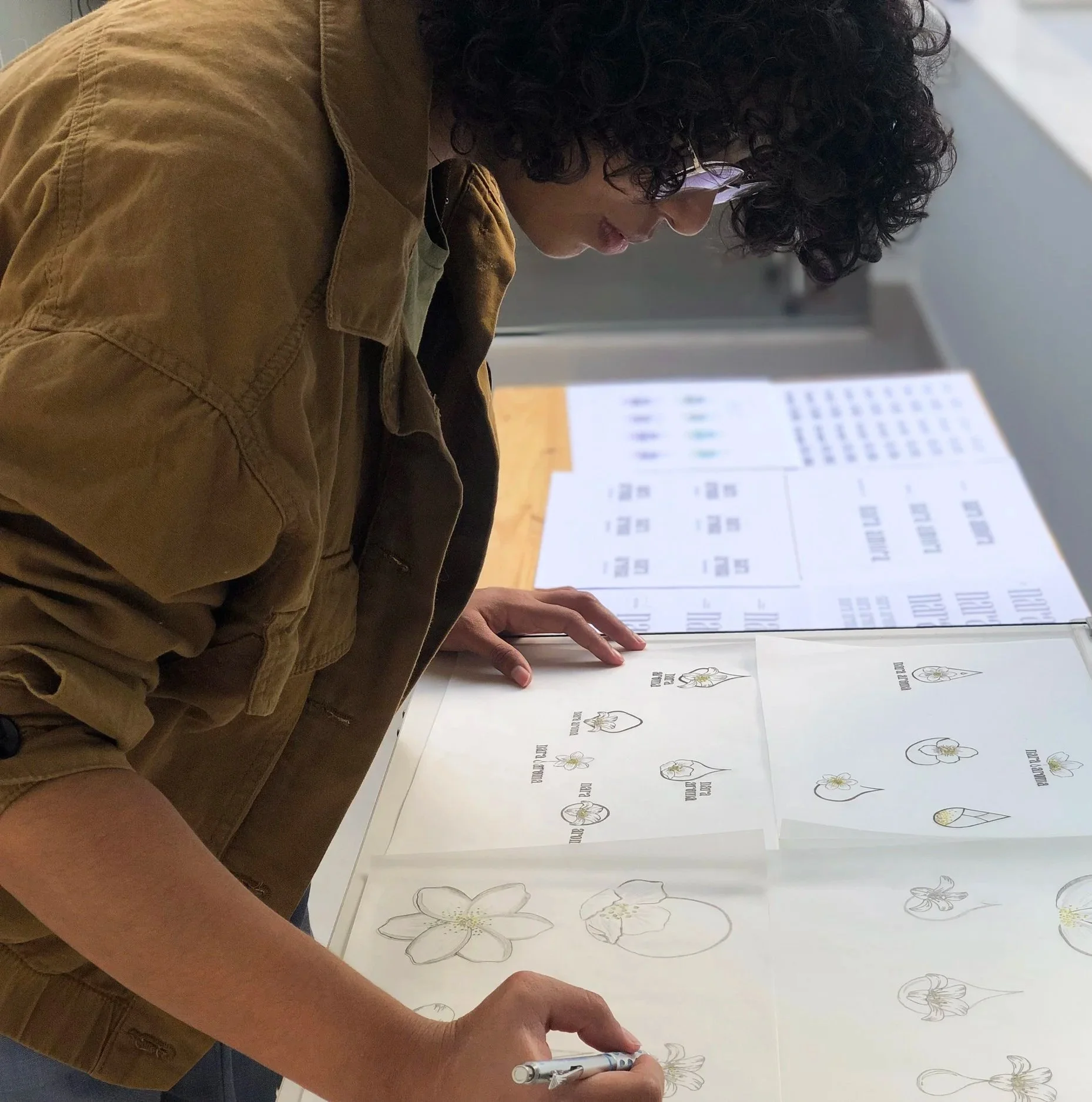 Pessoa com cabelo cacheado, óculos e jaqueta de algodão explorando desenhos de flores em um caderno em uma mesa.