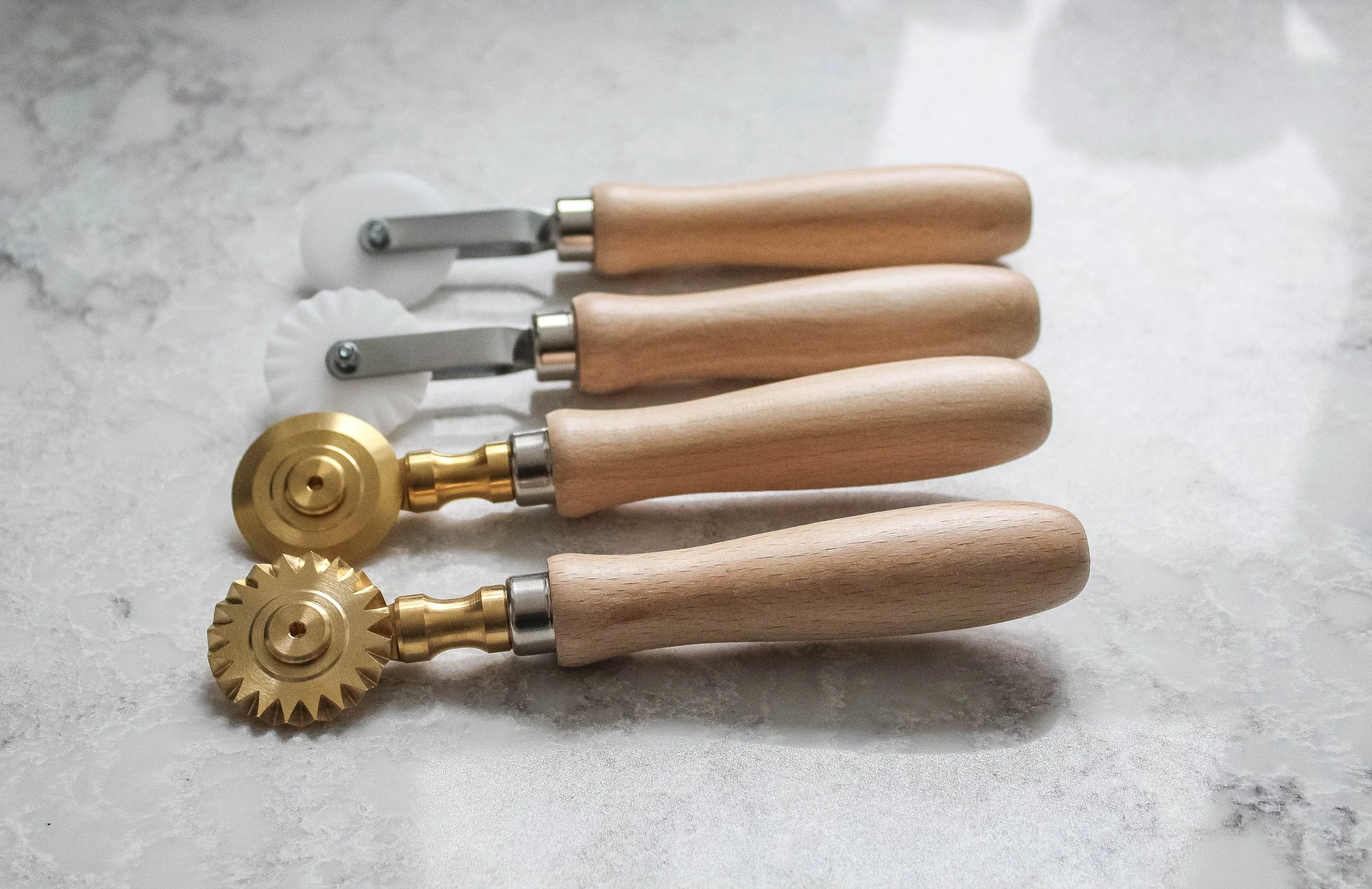 A set of pasta tools including four rolling cutters with wooden handles, two with fluted gold metal wheels and two with plain gold wheels, displayed on a white surface.