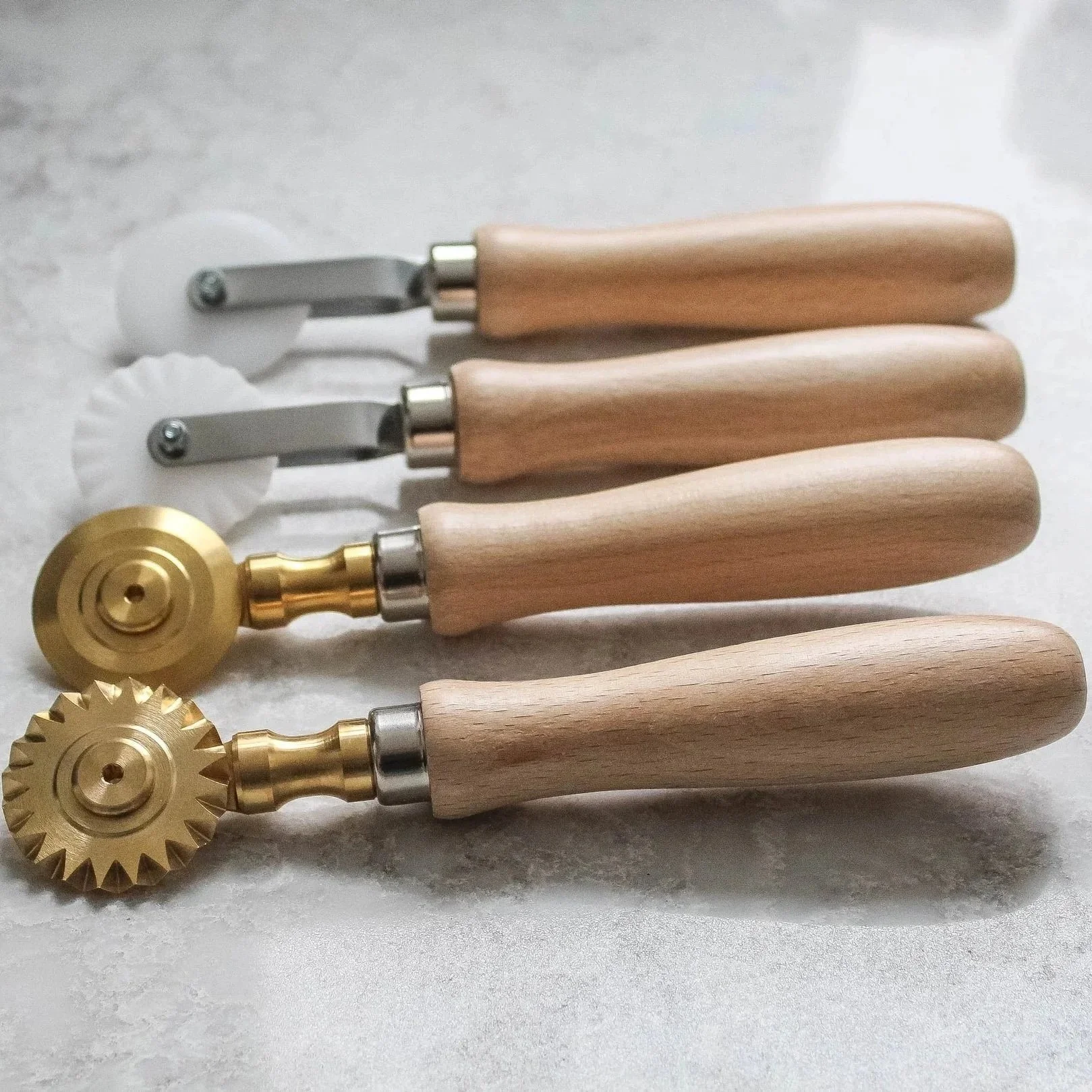 A set of pasta tools including four rolling cutters with wooden handles, two with fluted gold metal wheels and two with plain gold wheels, displayed on a white surface.