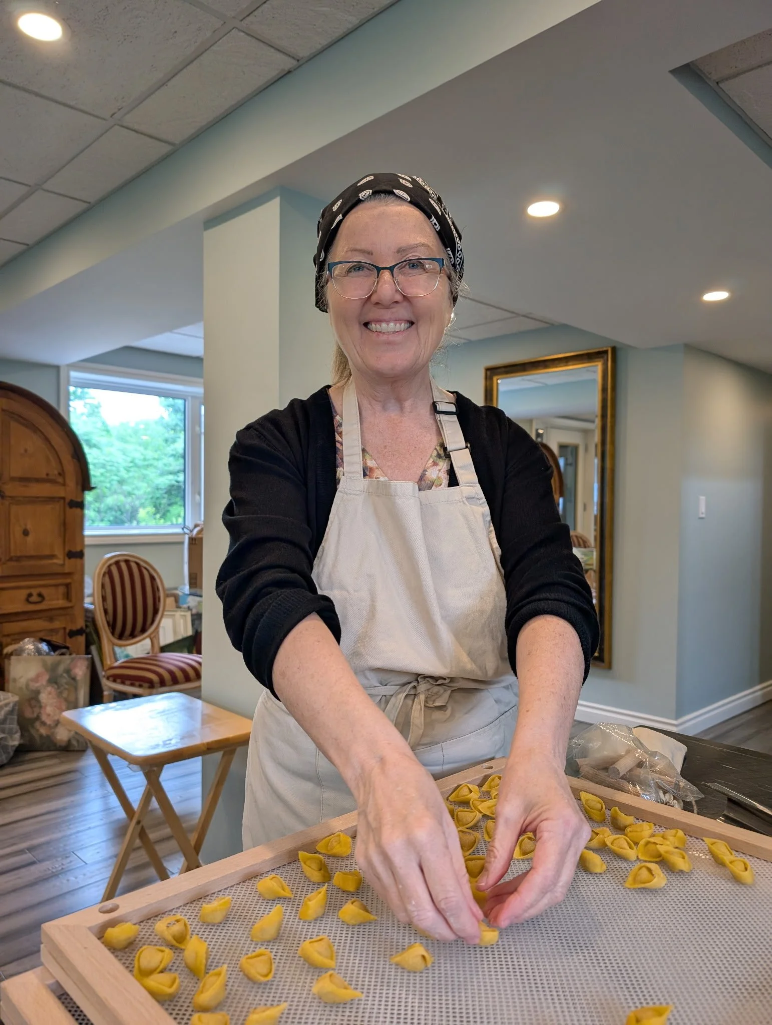 A woman with glasses and a black headscarf is smiling while making tortellini pasta in a bright room with wooden furniture and a window.