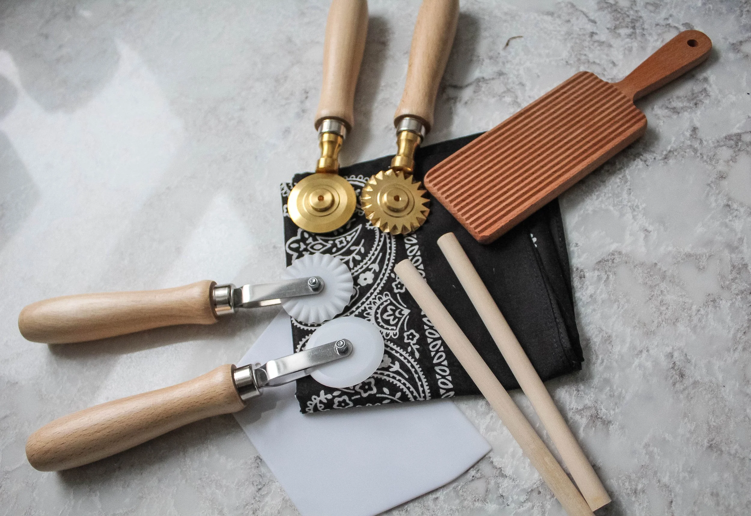Pasta Making tools including wooden dowels, pasta cutting wheels, bench scraper, wooden board, and a black black bandana on a light marble surface.