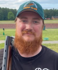 A young man with a red beard and mustache wearing a teal cap and black shirt, outdoors on a grassy field, smiling at the camera.