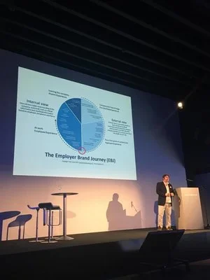 A person standing on stage giving a presentation about employee brand journey, with a large projected slide displaying a circular infographic and text, and a table with chairs and a small table on stage.