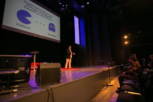 A person giving a presentation on stage in front of an audience in a dimly lit auditorium.
