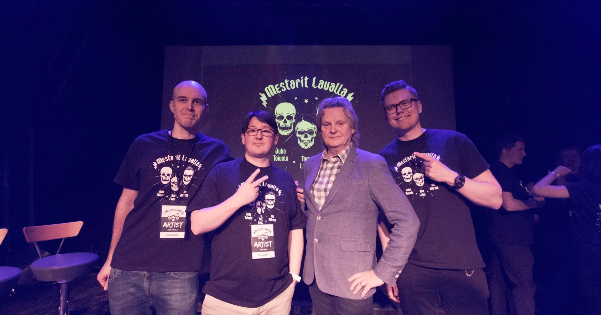 Four men standing together at an event, some wearing black shirts with skull graphics, one in a gray blazer, posing in front of a screen displaying skull graphics and the text 'Mestorit Lavulla'.