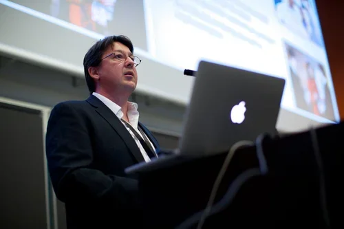 A man in a suit giving a presentation with a MacBook in front of him and a projection screen behind him.