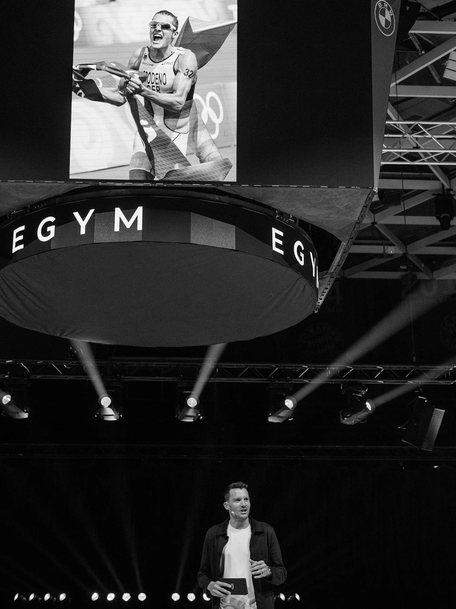 Jan Frodeno speaking on stage at an event, with a large screen behind him displaying a photo of him winning the 2008 Olympic Gold Medal in Peking.