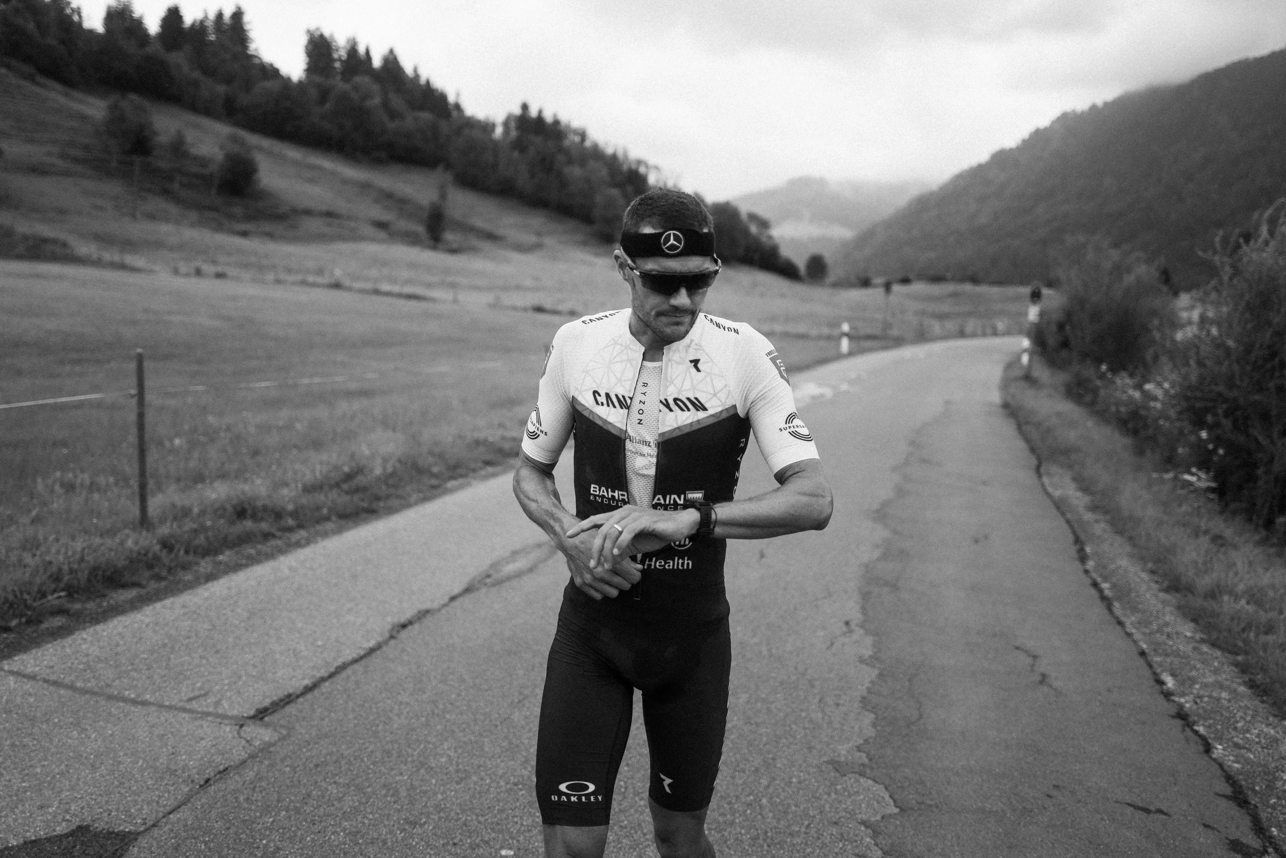 Jan Frodeno stands on a rural road, looking at his watch, with a mountainous landscape in the background, in black and white.