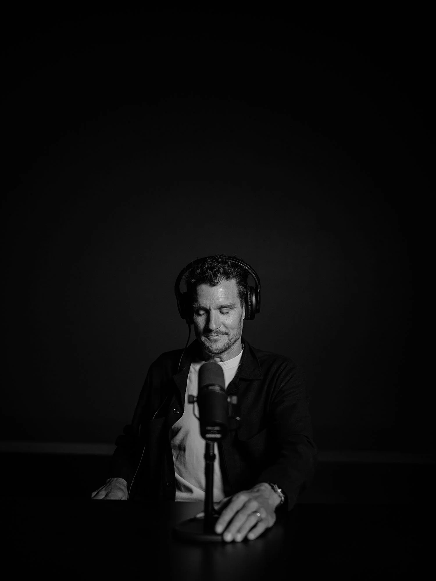 Black and white photo of Jan Frodeno wearing headphones sitting at a table with a microphone, looking down with a smile.