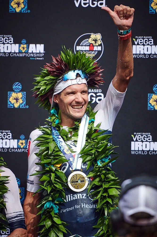 Jan Frodeno wearing a wreath of leaves, sunglasses, and a white headband, celebrating on a podium with a medal around his neck at the Ironman World Championship in Hawaii.