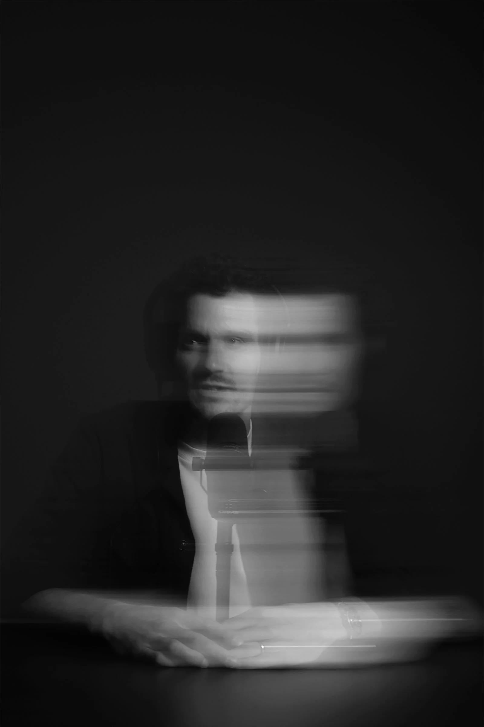 A black and white photo of Jan Frodeno sitting at a table, holding a smartphone, with a camera in front of him. The image has motion blur. The background is dark.