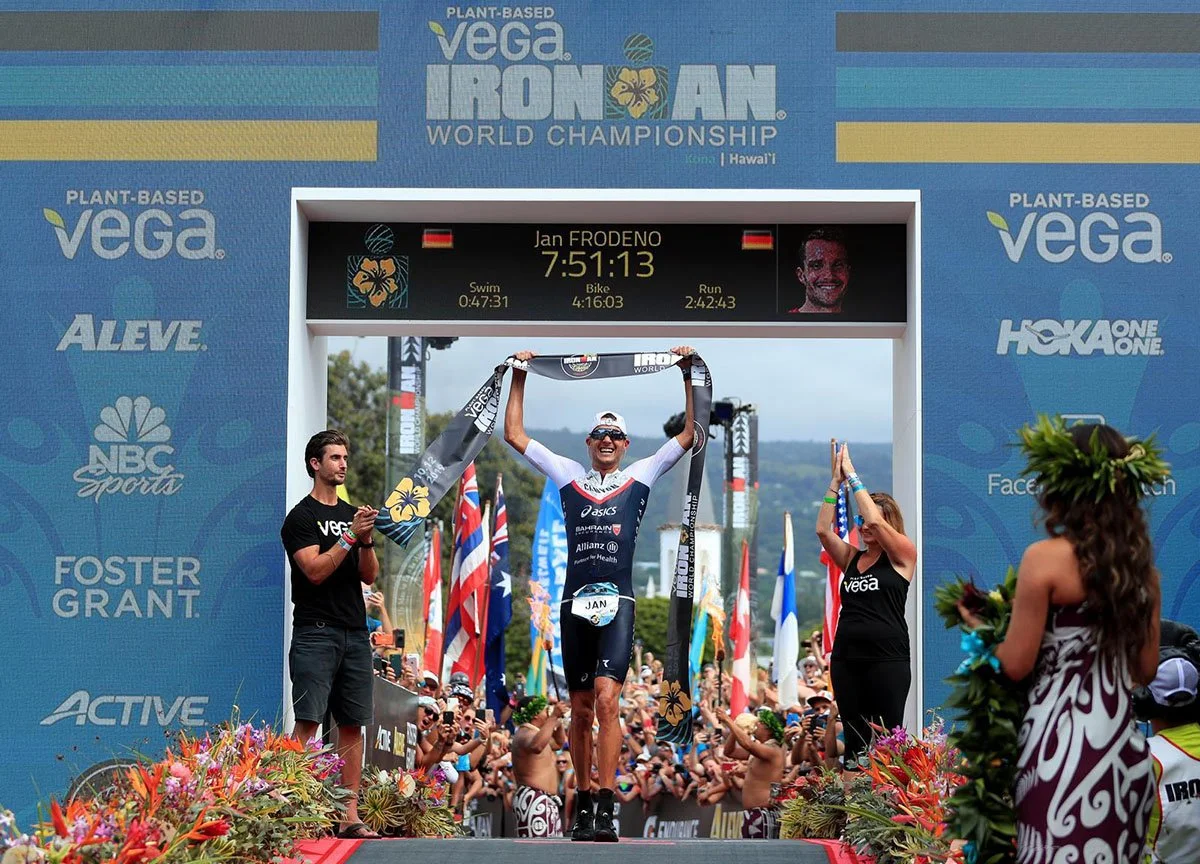Jan Frodeno crossing the finish line, smiling and raising arms with a medal, crowd cheering, flags, and event branding for the VEGA Ironman World Championship in Hawaii.